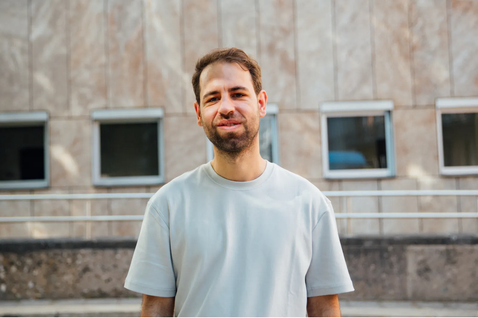 Alexander Ali Gutsfeld trägt ein helles T-Shirt und steht draußen vor einem Haus mit vielen Fenstern.