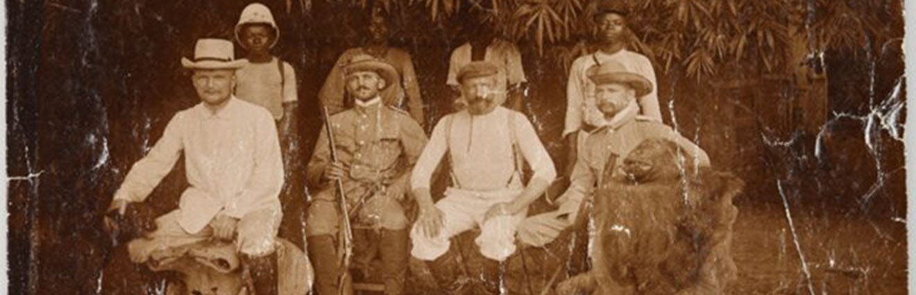 Männer in Strohhüten umringt von Offizieren der Kaiserlichen Schutztruppe in Kamerun, rechts ein Affe