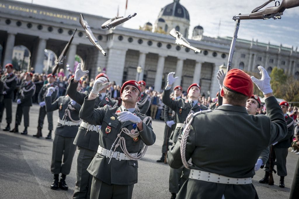 Das Foto zeigt die Heeres-Schau am Nationalfeiertag in Wien: Soldat:innen des Österreichischen Bundesheers mit roten Baretten werfen bei einer Vorführung ihre Gewehre in die Luft. Im Hintergrund sieht man historische Gebäude am Heldenplatz.