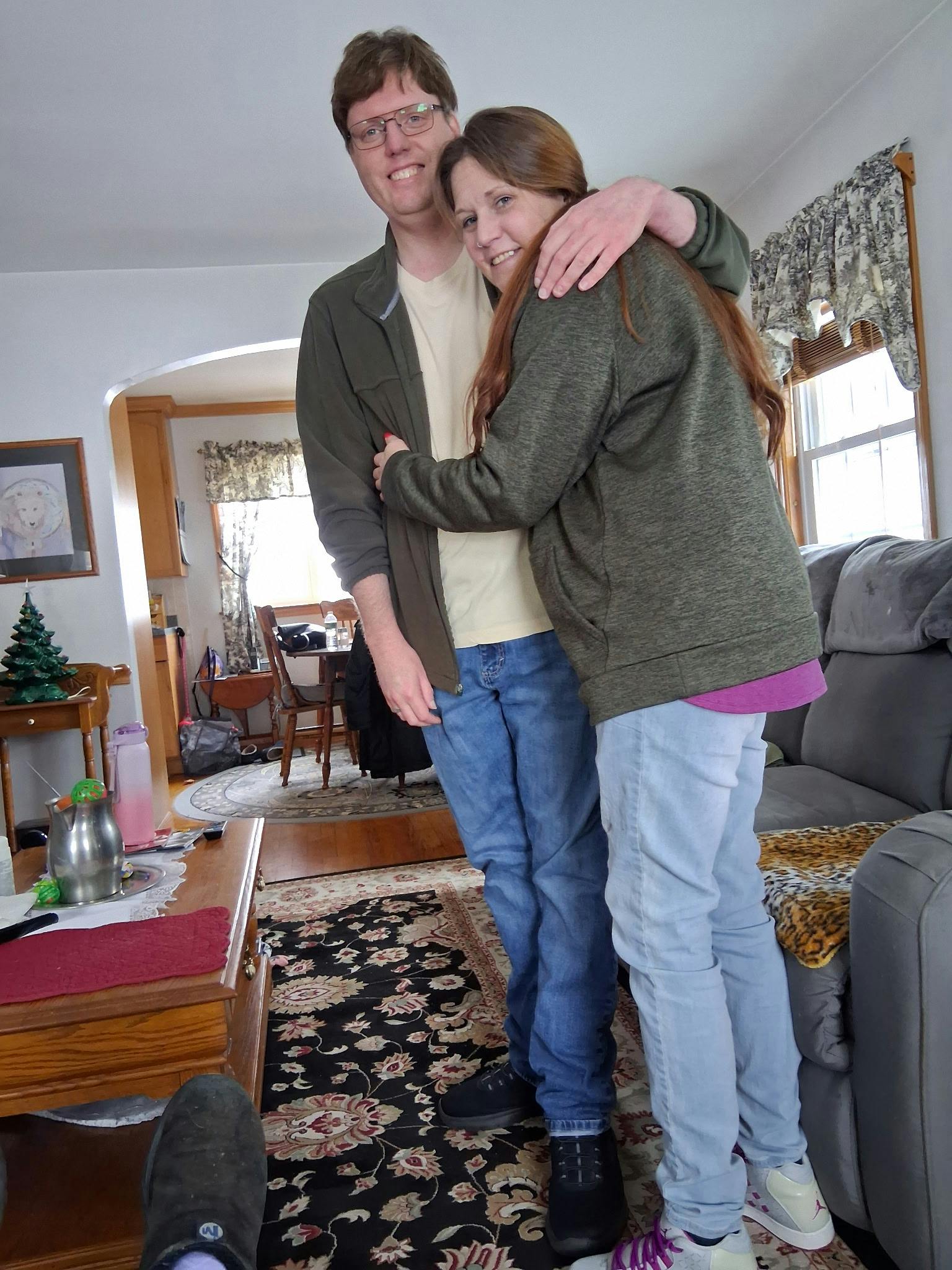 Danielle and I hugging in January of 2026. I'm on the left and she's on the right. My face is a little weird because I had to explain to the "photographer" how to use a smartphone. I'm wearing jeans, a yellow T-shirt, and a green sweatshirt. I have black framed glasses on my face. Danielle is wearing jeans, a purple shirt and grey sweatshirt. A coffee table is on the left and a grey couch on the right. We are standing on a black and white floral patterned rug.