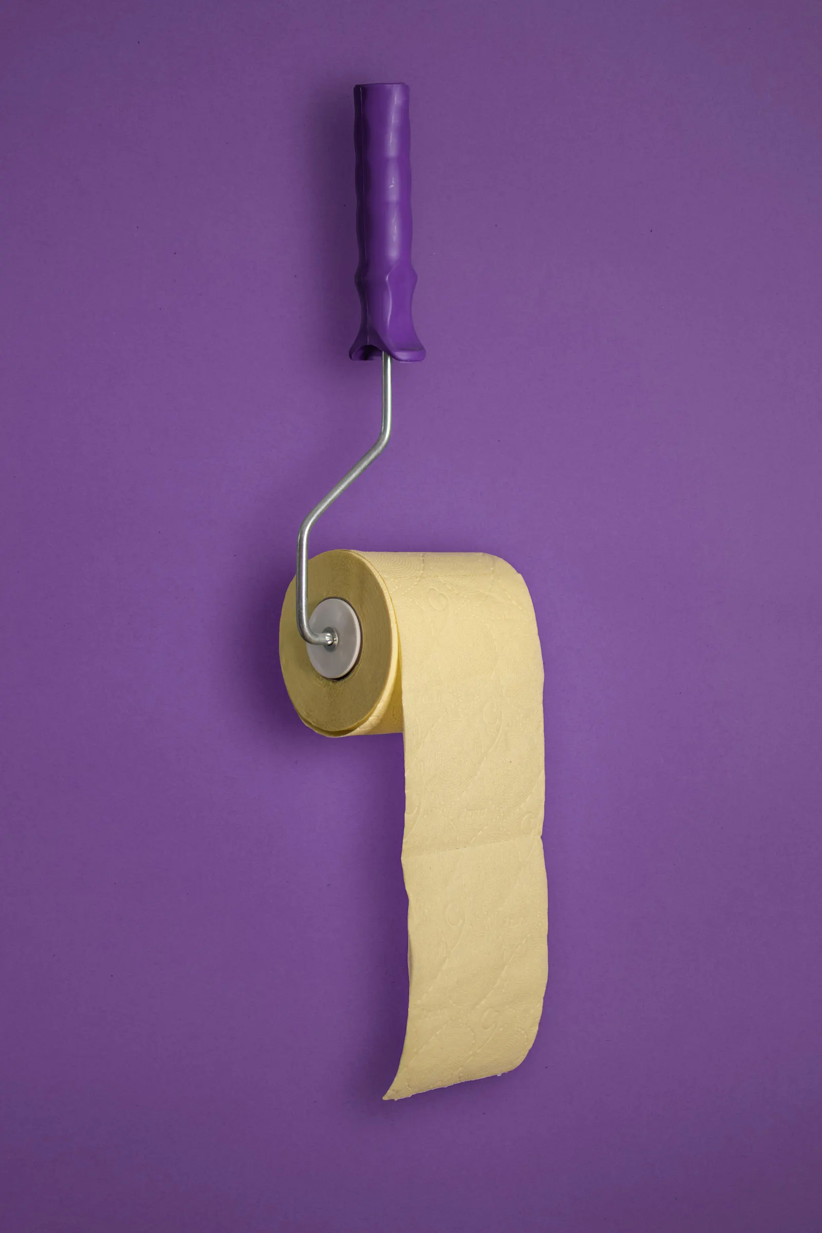 Lila Hintergrund. Davor eine lilafarbene Malrolle, an der eine Rolle gelbes Toilettenpapier hängt.