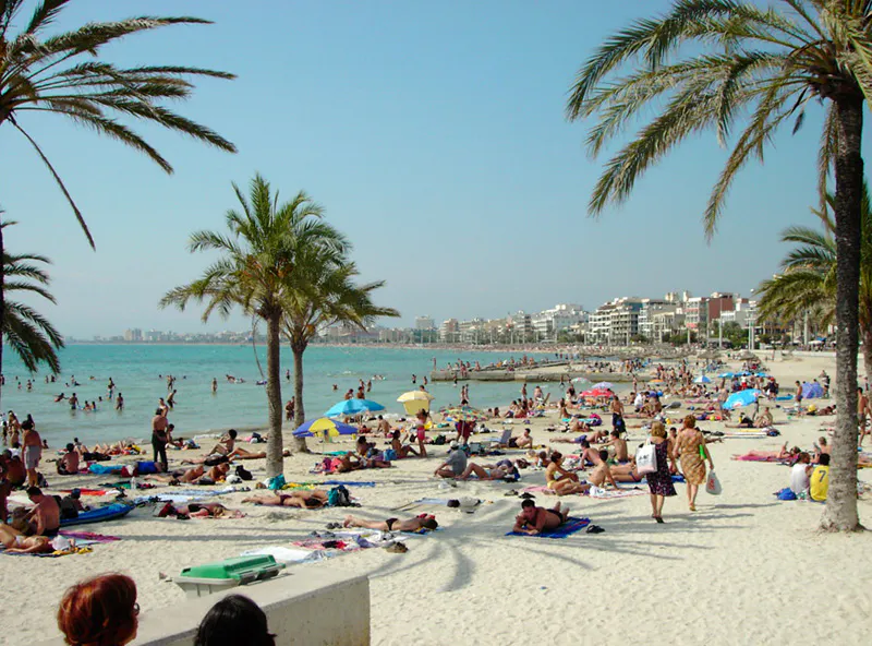 Historische Aufnahme eines Strands auf Mallorca. Im Hintergrund Häuserblocks, im Vordergrund weißer Sand mit Palmen und Besucher.