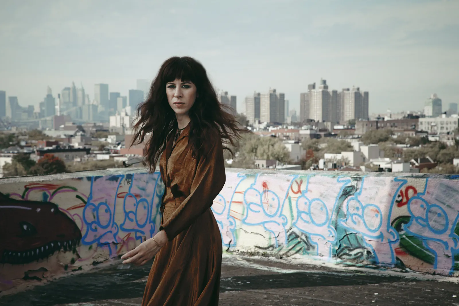 Auf dem Bild sieht man im Vordergrund die Komponistin Missy Mazzoli. Sie trägt ein langes, braun-goldenes Kleid mit einem feinen Muster und schaut mit ernstem, leicht nachdenklichem Blick in die Kamera. Ihr dunkles, welliges Haar fällt locker über ihre Schultern.
Sie steht auf einem Dach, das mit Graffiti bemalt ist – auffällig sind mehrere bunte Figuren an einer niedrigen Wand hinter ihr.
Im Hintergrund erstreckt sich ein Teil der Skyline von New York City. Die charakteristischen Hochhäuser und die verschwommen sichtbare Brooklyn Bridge deuten auf Manhattan hin. Der Kontrast zwischen der rauen Street-Art-Ästhetik und der Eleganz von Mazzolis Erscheinung erzeugt ein spannungsvolles, künstlerisch aufgeladenes Bild.