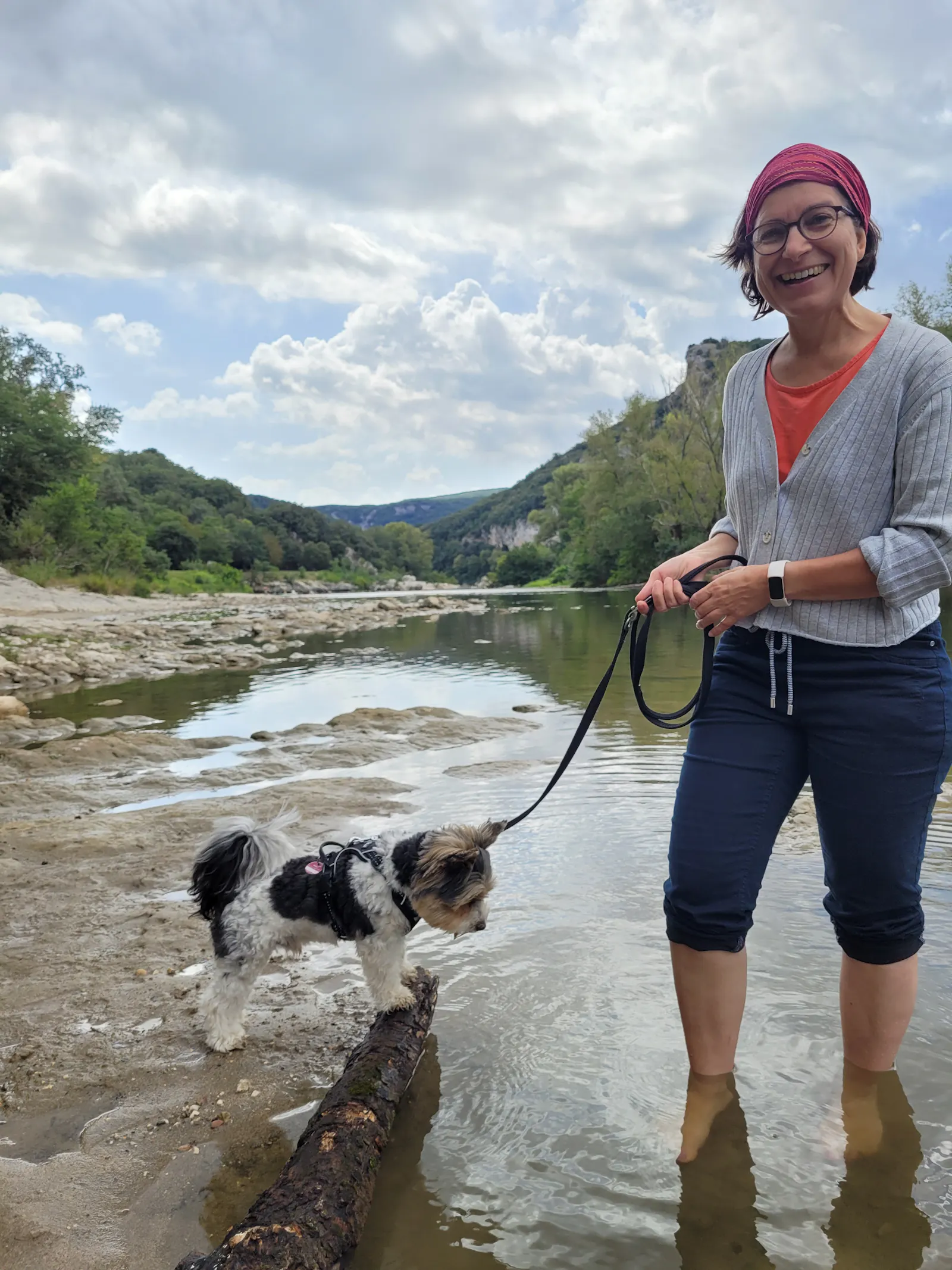 Frau steht mit den Füßen in einem seichten Flussbett, hält einen kleinen Hund an der Leine, der auf einem Felsen steht.