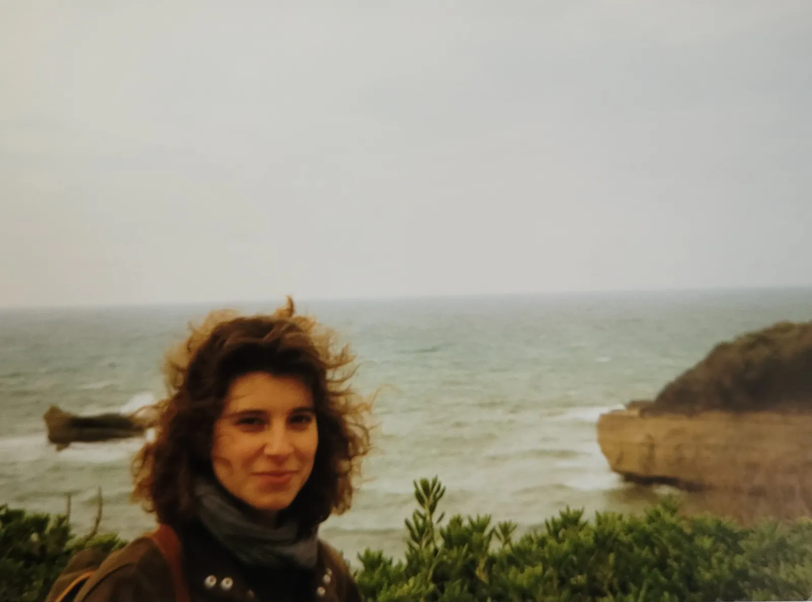Junge Frau mit langen wehenden Haaren am Strand von Biarritz, im Hintergrund Felsen, der Himmel ist grau.