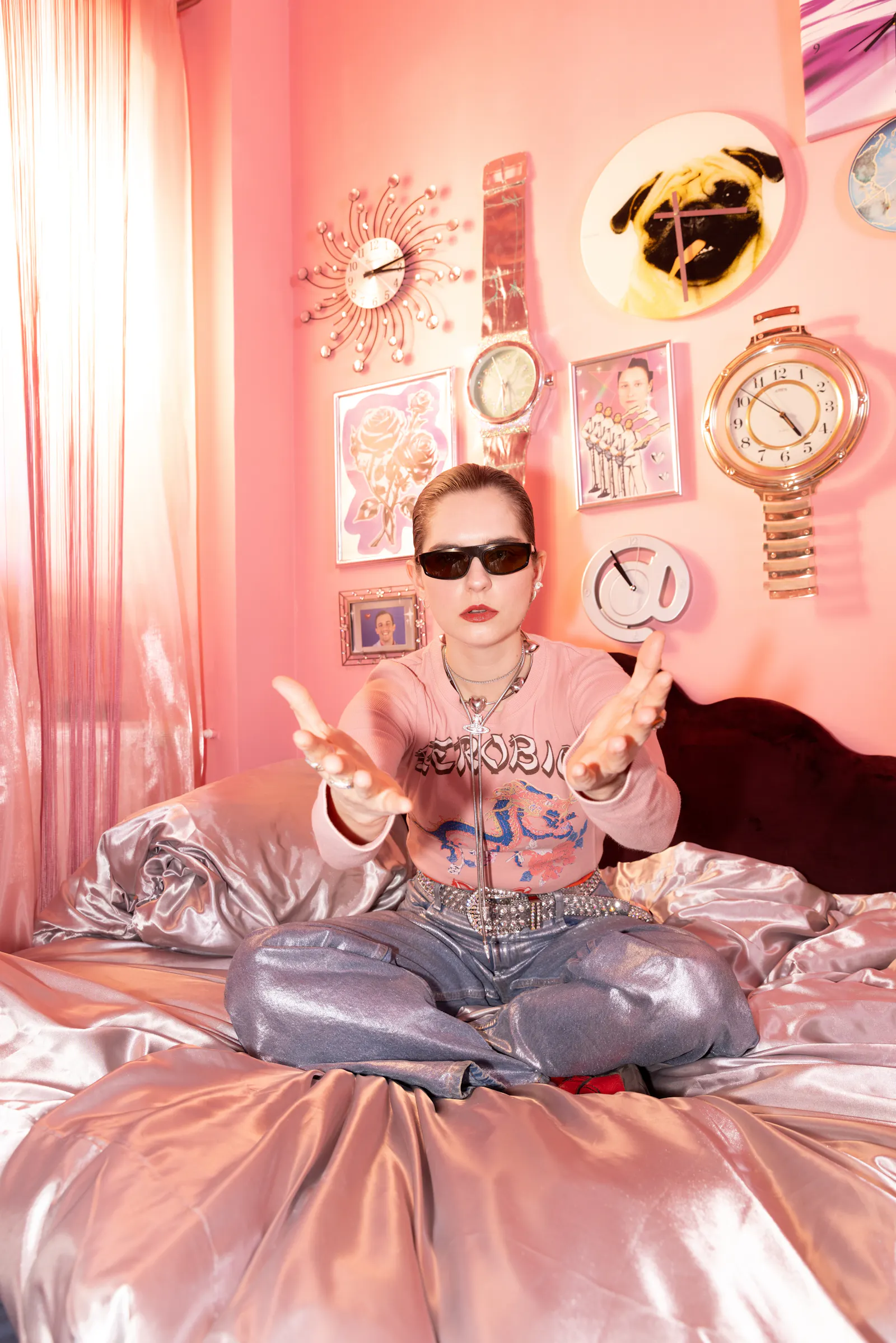 Foto der Musikerin Fuffifufzich, die im Schneidersitz und mit Sonnenbrille auf einem Bett sitzt, die Hände in richtung Kamera ausbreitend, im Hnitergrund diverse Souvenir-Uhren an der Wand, alles in rosa Farben gehalten