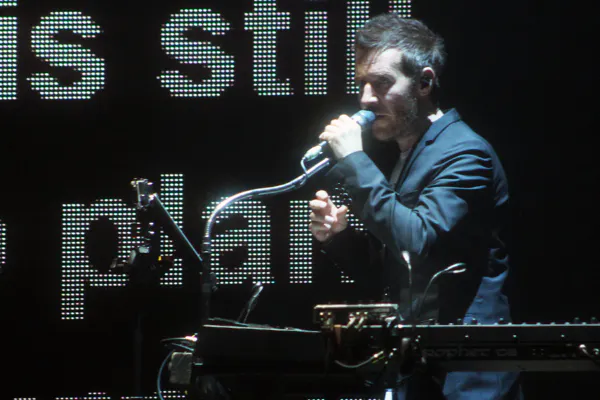 Foto des Musikers Robert Del naja auf einer Bühne mt einem Mikrofon und Keyboards vor einer Video/LED Wand mit Buchstaben