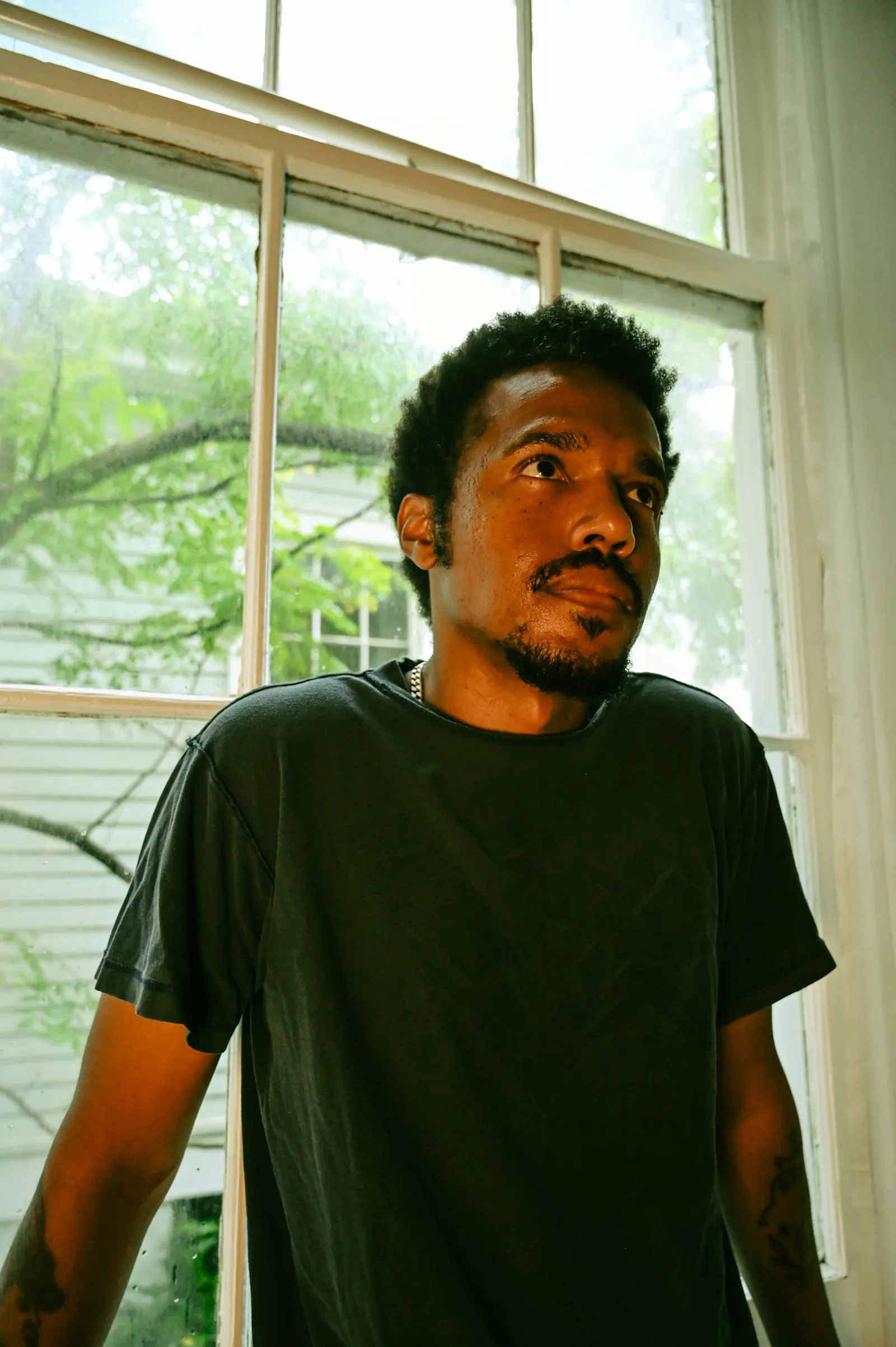 Foto des Musikers Benjamin Booker, der mit dem Rücken vor einem Fenster mit Blick in einen Baum steht und an der Kamera vorbei in die Ferne guckt