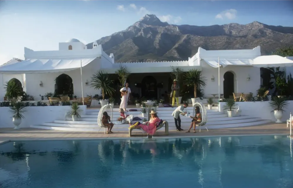Im Hintergrund Berge und eine weiße Villa, im Vordergrund sitzen Menschen an einem großen Pool
