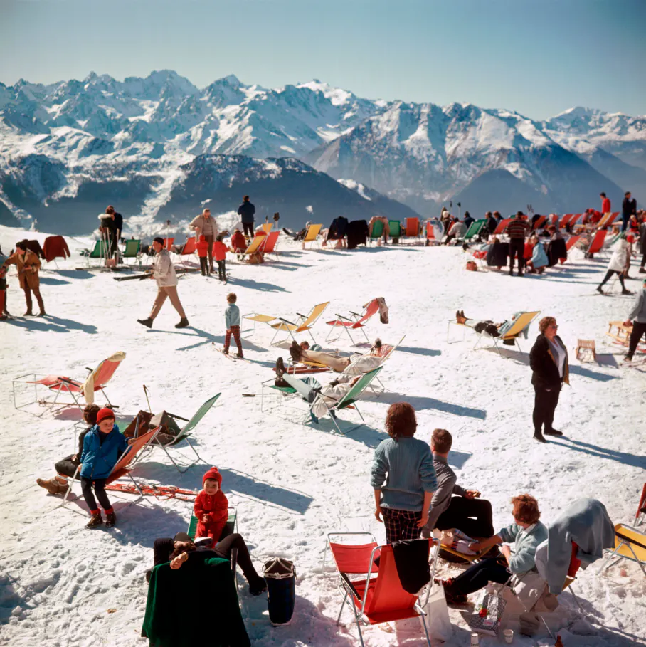 Im Vordergrund Menschen auf Sonnenstühlen im Schnee, im Hintergrund Berge