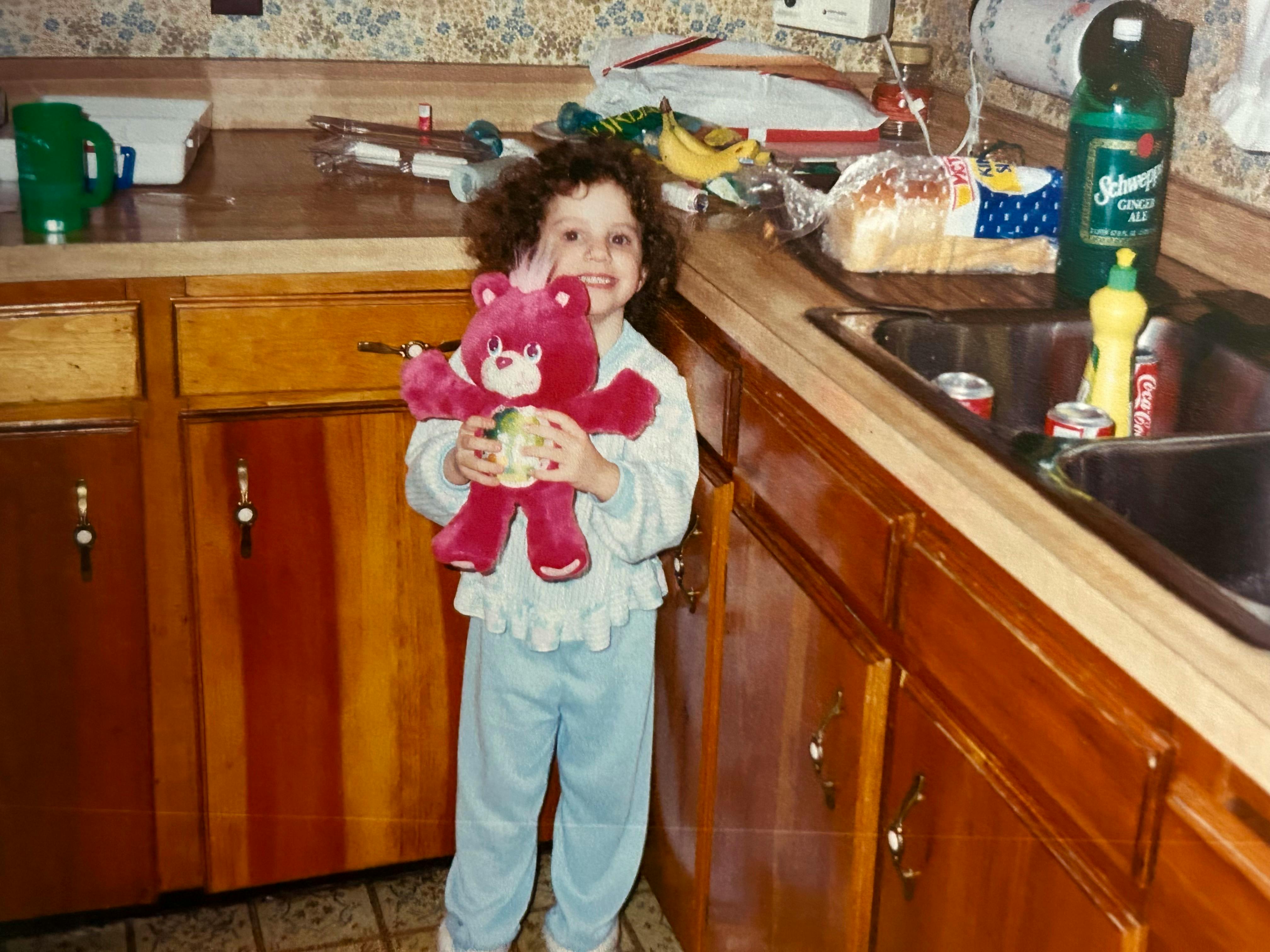 Hannah as a toddler standing in the old kitchen and proudly showing off her new red Care Bear. The counters are worn and covered in cups, a loaf of Wonder bread, bananas and asthma treatment tools. Bottles of ginger ale and coke are around and in the sink on the right along with a yellow bottle of dish soap.