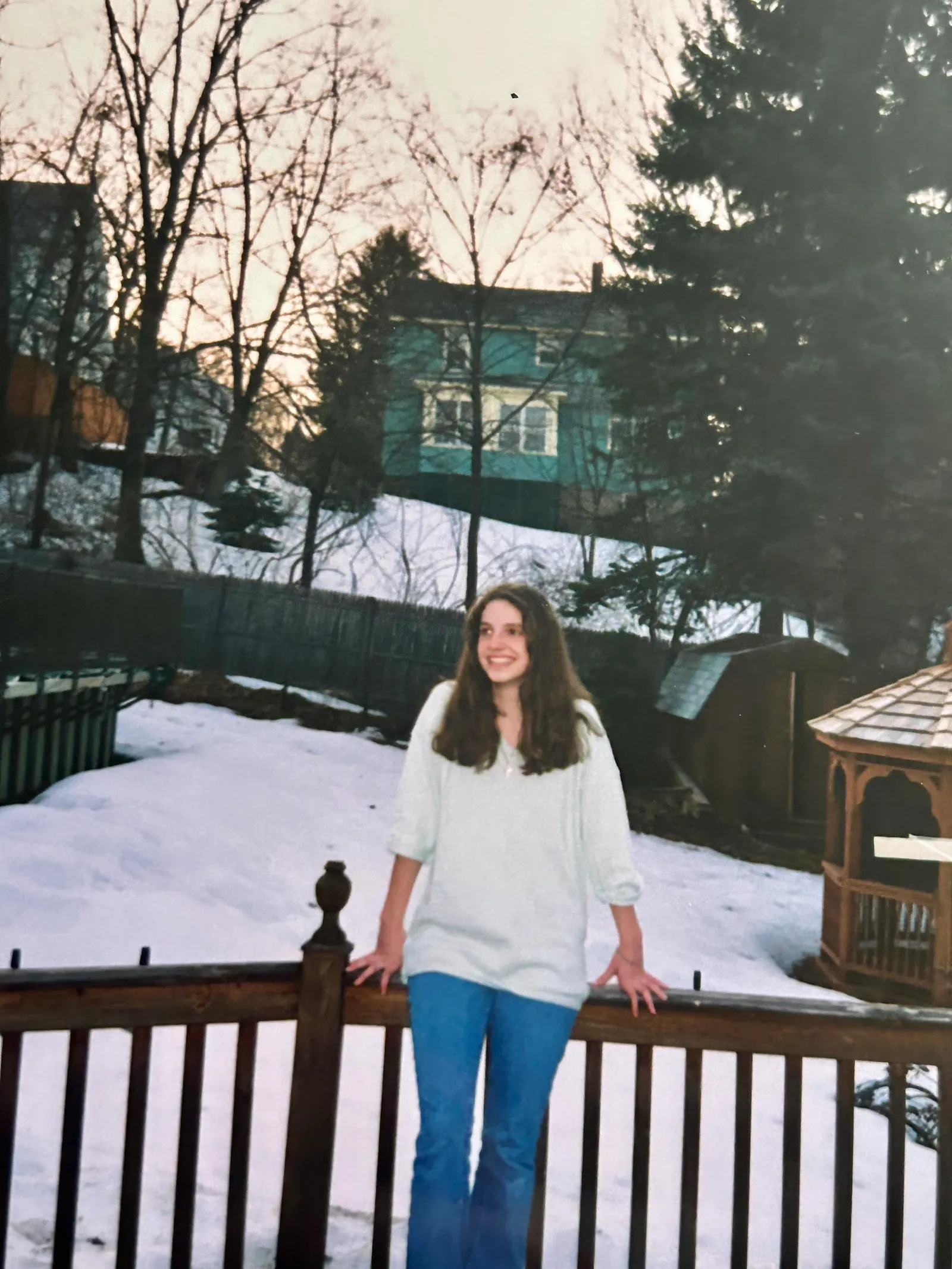 A teenage Hannah dressed in a white sweatshirt and jeans poses on the edge of our deck in winter. There is snow on the ground. A gazebo is in the background on the right. An above ground pool is on the left. A neighbouring green house is in the background.