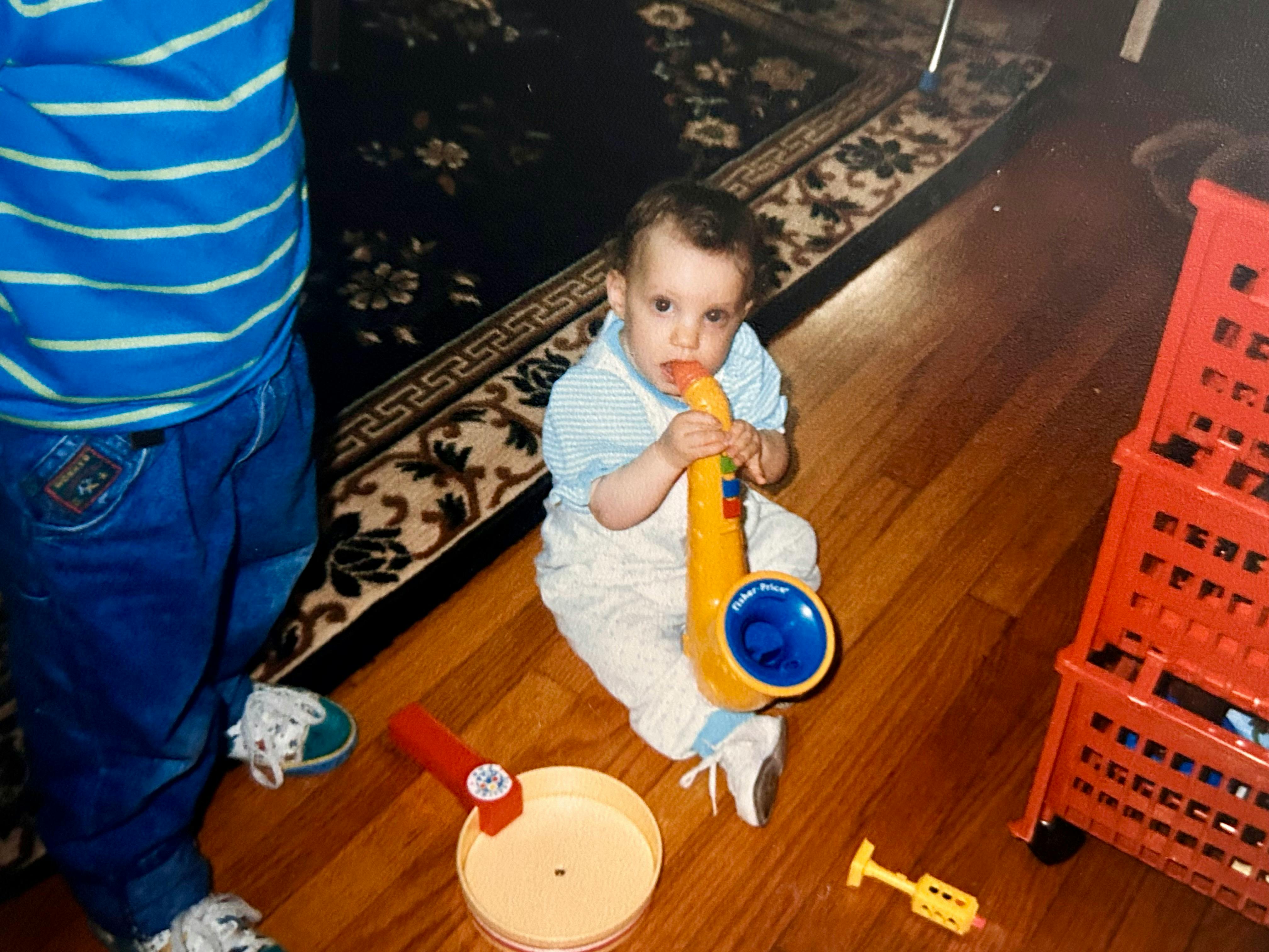 A baby Hannah playing with a yellow saxophone toy on the floor. A red cart of toys and crafts is to the right. You can see my torso on the left in a blue and white shirt as I watch her play. A toy pan is between us. A black rug with white floral patterns is in the background.