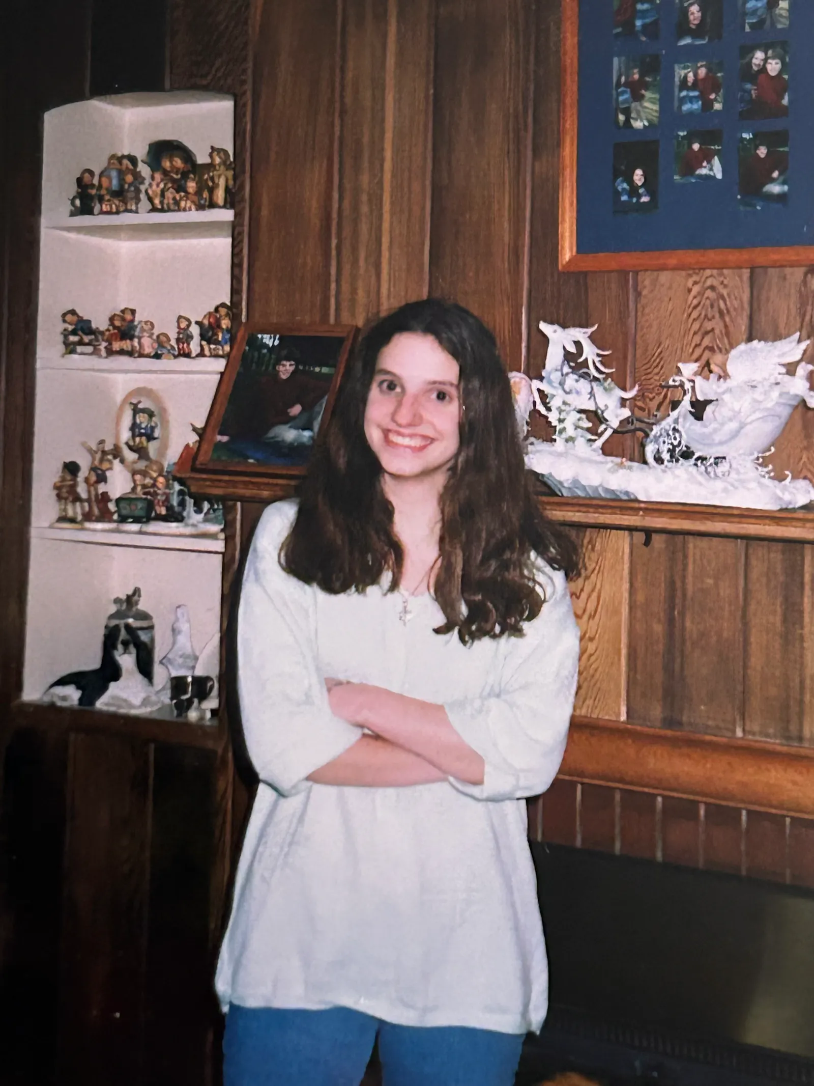 A teenage Hannah dressed in a white sweatshirt and jeans poses before the fireplace and mantle. Our pictures are above the mantle while a white sleigh pulled by a white reindeer decorates the middle of it. She has her arms crossed and is smiling.