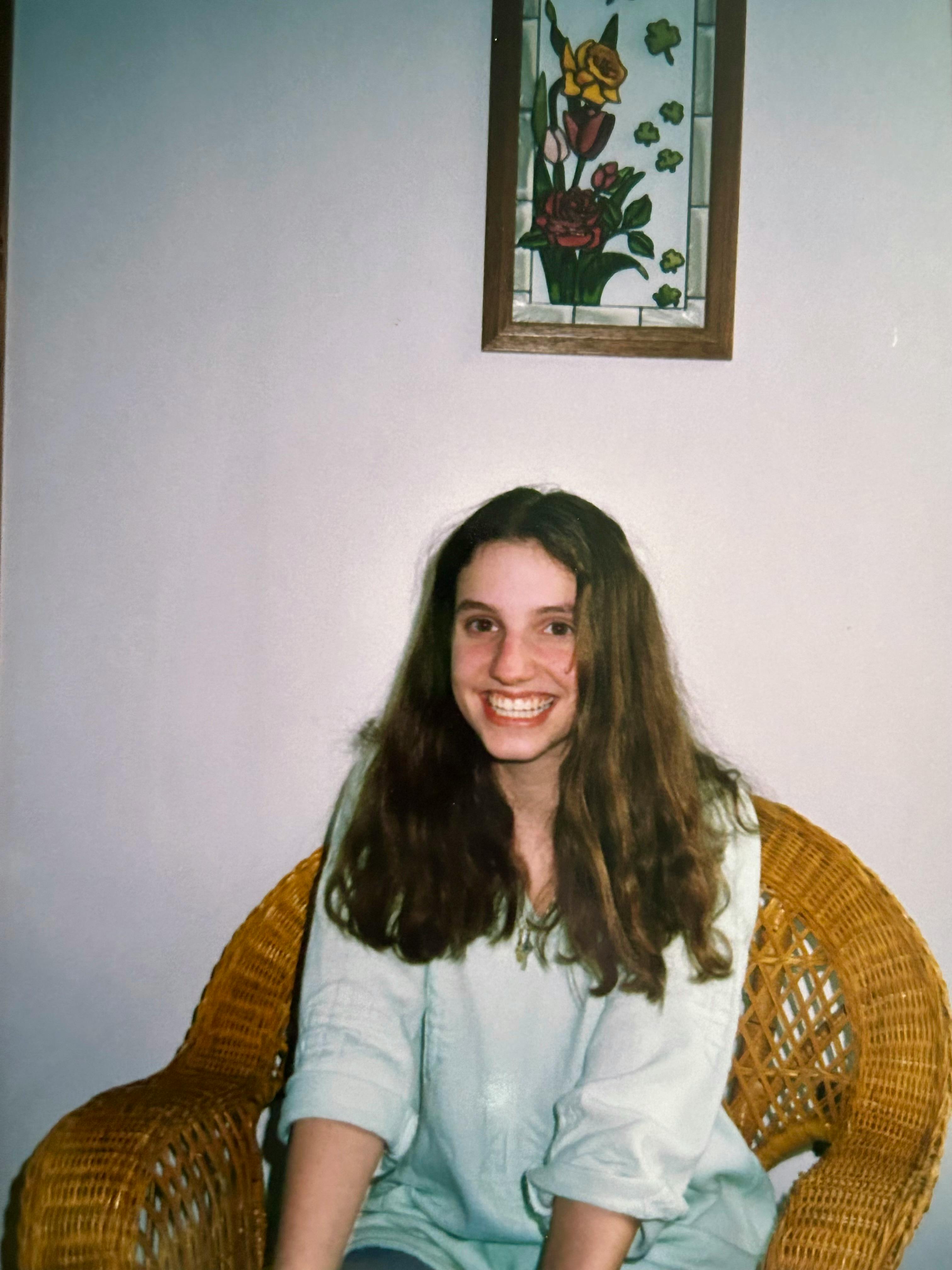 A teenage Hannah dressed in a white sweatshirt and jeans poses in a brown wicker chair beneath a stained glass artwork showing a bouquet of red, pink, and yellow roses.