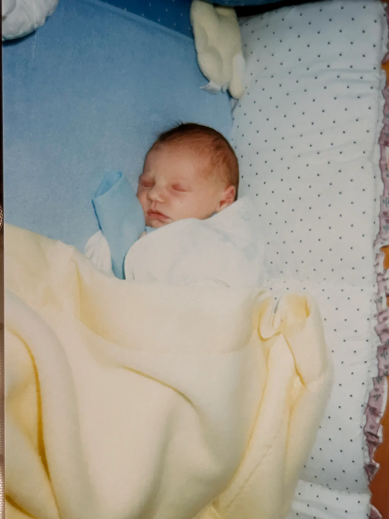 A newborn Hannah in her crib under a yellow blanket and atop a blue bedding. A white cushion with blue dots is on the right of the crib to prevent her from falling out.