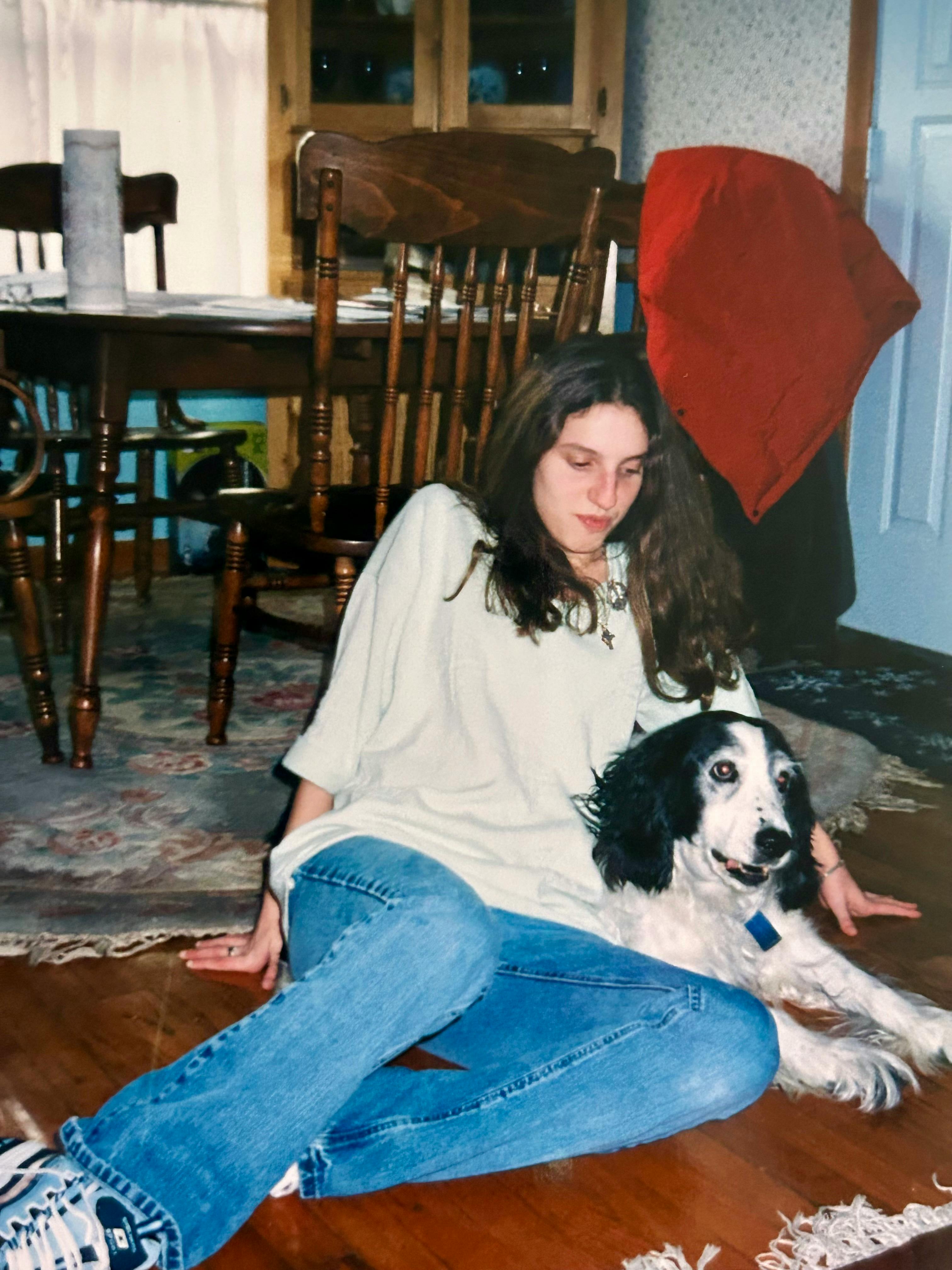 A teenage Hannah dressed in a white sweatshirt and jeans sits on the floor with our old dog Jackson, a black and white springer spaniel, with the dining room table behind them both. A red placemat is draped over a chair on the right. The dining room table is behind them both atop a green and pink rug.