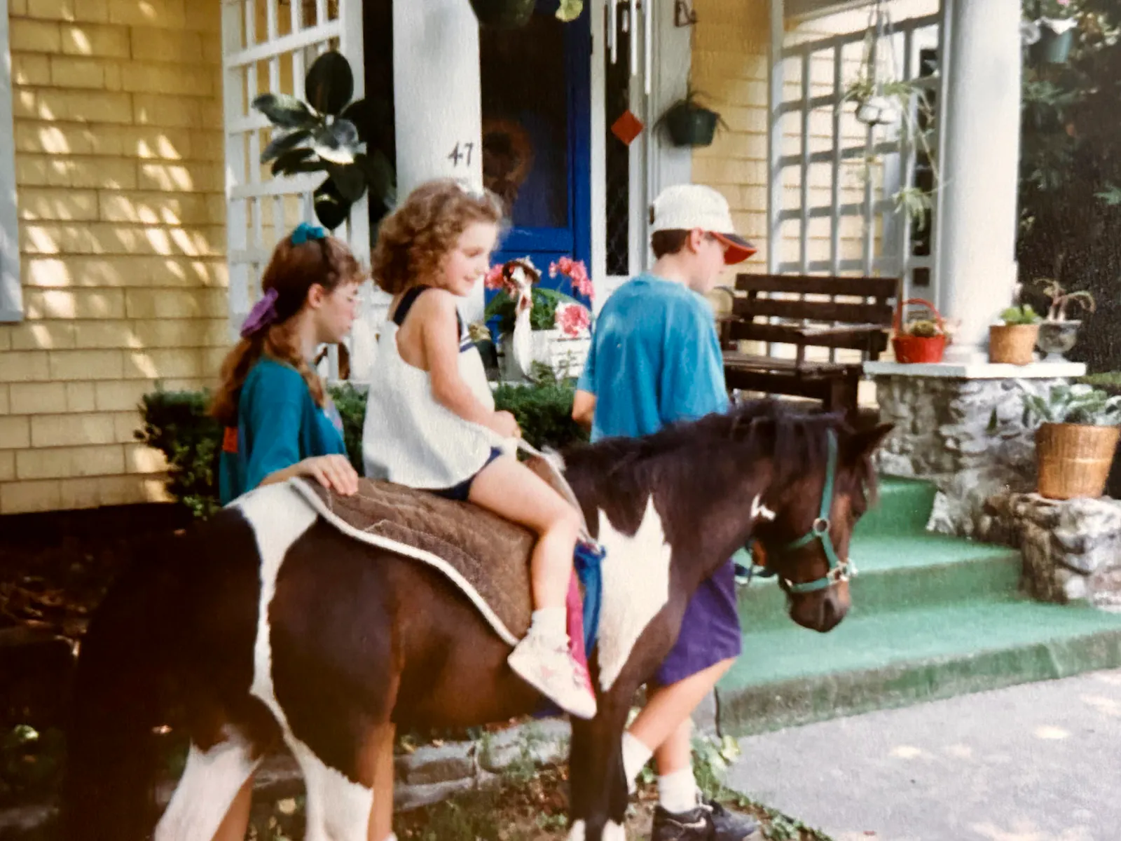 Hannah takes a pony ride at a local petting zoo. She sits atop the brown and white pony and smiles in her white dress. Two teenagers dressed in blue uniform shirts are on the side to guide the horse on its path. A yellow building is in the background with a blue door for an entrance.