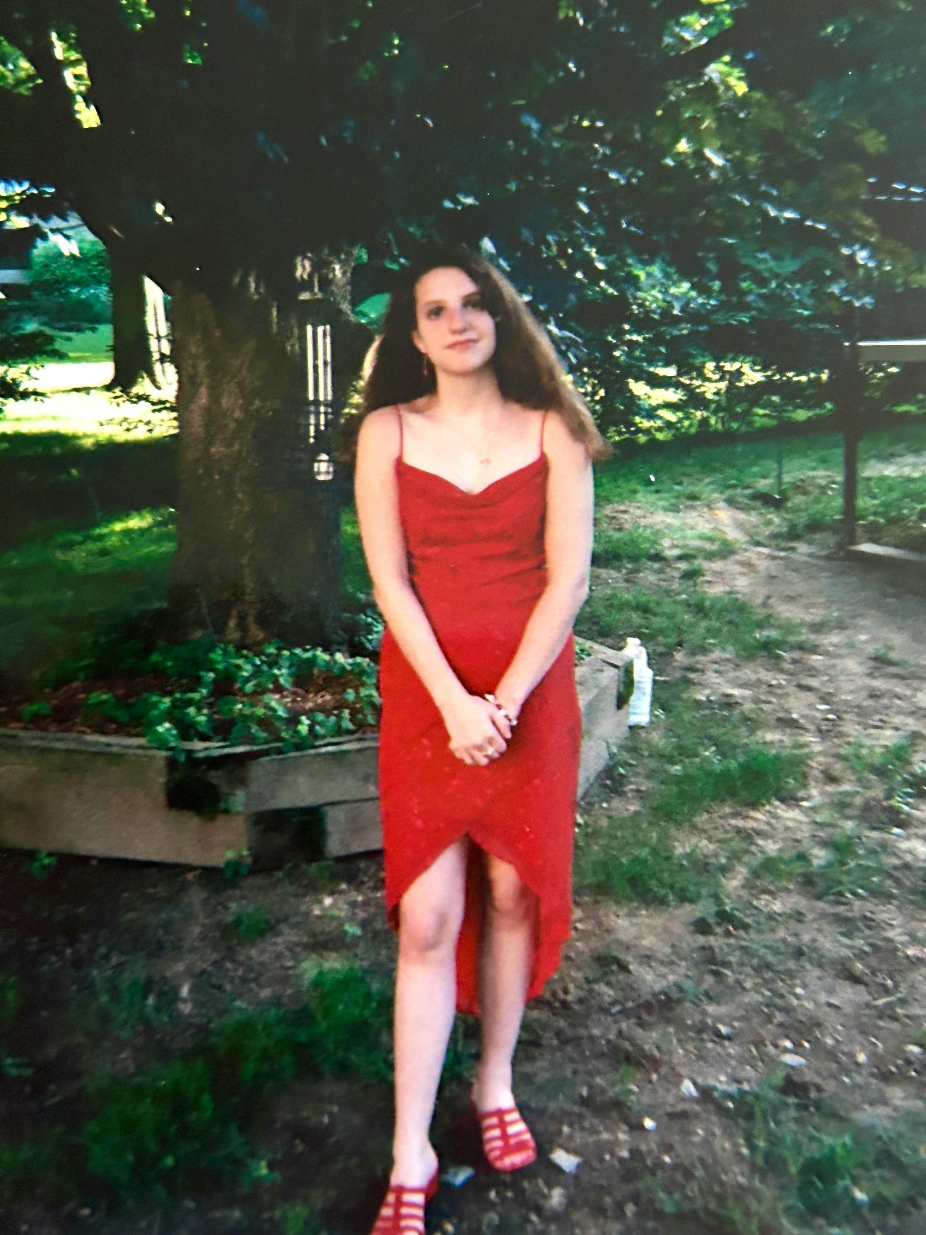 Hannah in a red dress and her hair left loose as she prepares to graduate from the 8th grade. She stands beneath a red maple tree and before a garden with green plants beginning to spring to life. The grass is spotty with large, bare patches of dirt between them. The sun is beginning to set in the distant background.