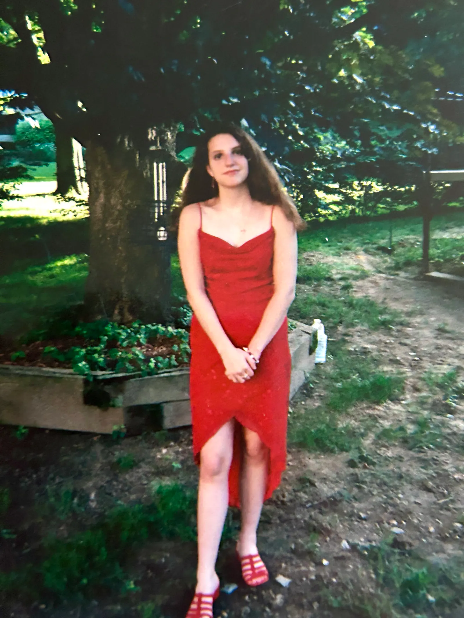 Hannah in a red dress and her hair left loose as she prepares to graduate from the 8th grade. She stands beneath a red maple tree and before a garden with green plants beginning to spring to life. The grass is spotty with large, bare patches of dirt between them. The sun is beginning to set in the distant background.