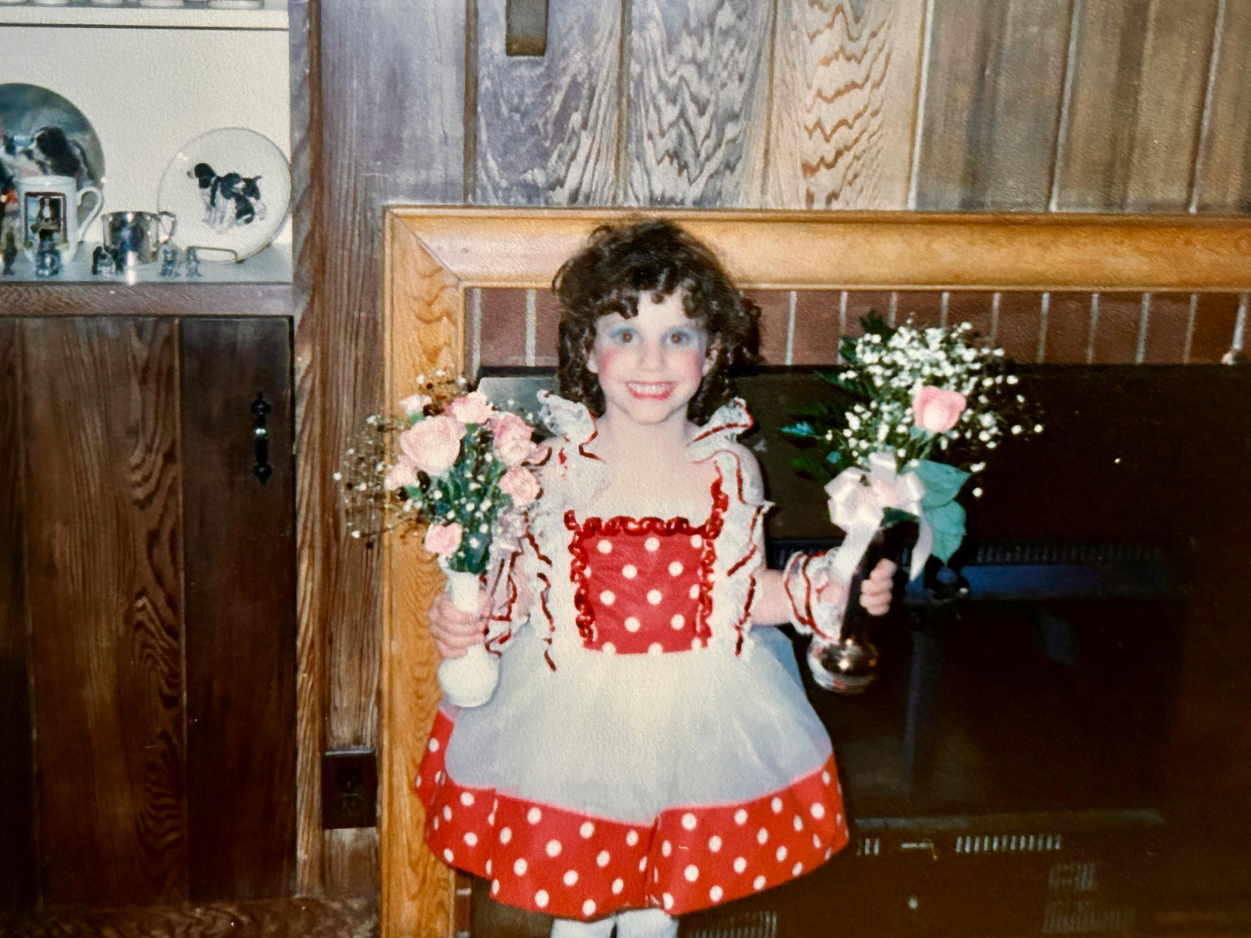 Hannah dressed for her first ever dance recital. She wears exaggerated makeup (blue around her eyes, and red cheeks) while wearing a polka dot dress. She holds a bouquet of pink and white flowers in both hands. The shelves in the background are adorned with figures and plates showing black and white springer spaniels. A black fireplace is behind her.