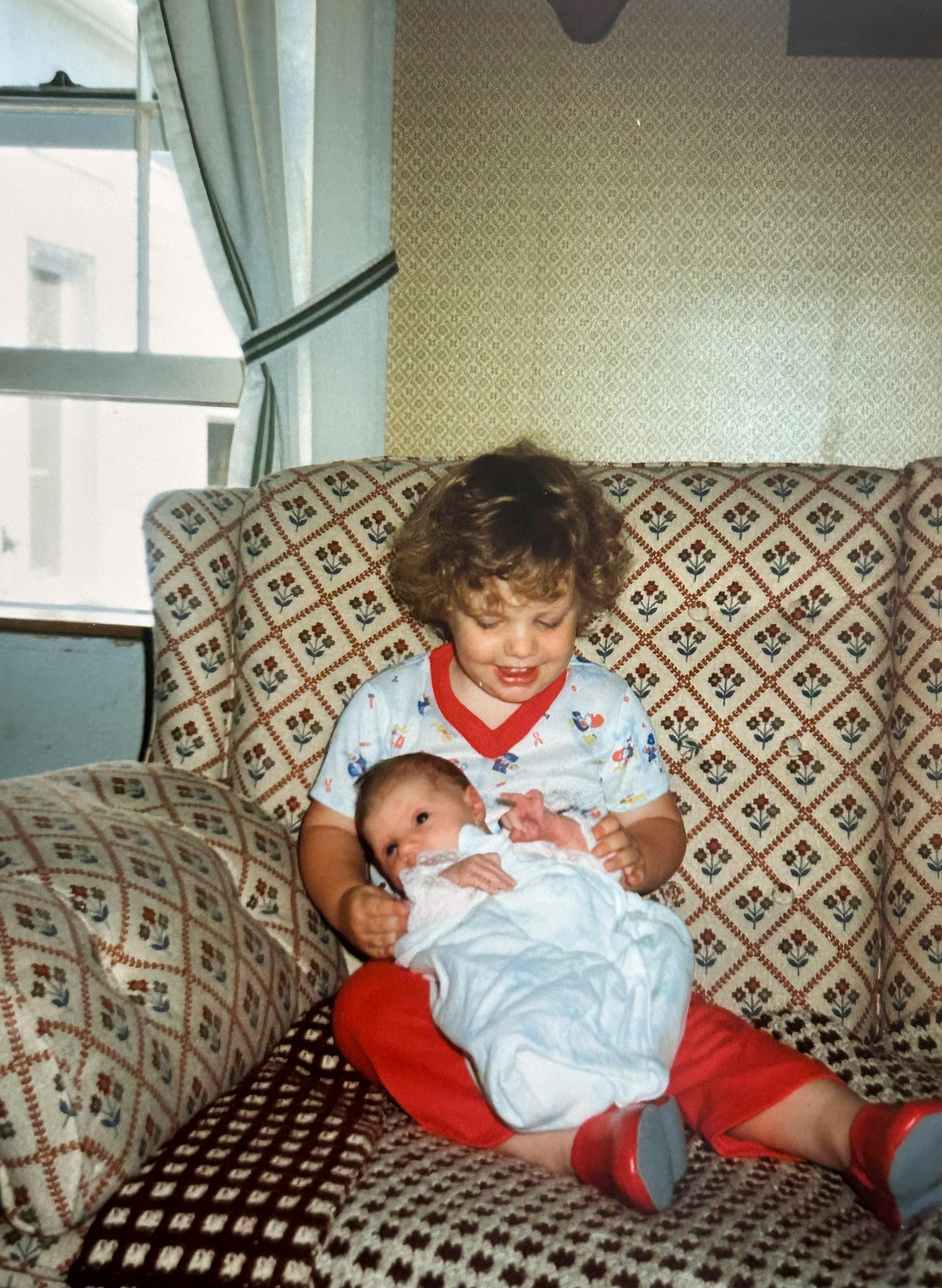 A month old Hannah sitting in my 2-year-old lap. She appears to be trying to sit up while wrapped in a blue blanket. I’m wearing a white outfit with red edges and gaudy red pants. We are  sitting on a brown afghan atop a brown couch with dark brown, red, and blue flower patterns. The greenish/brown wallpaper has similar diamond patterns upon it. Behind the couch is a window with white and green curtains. Some style!