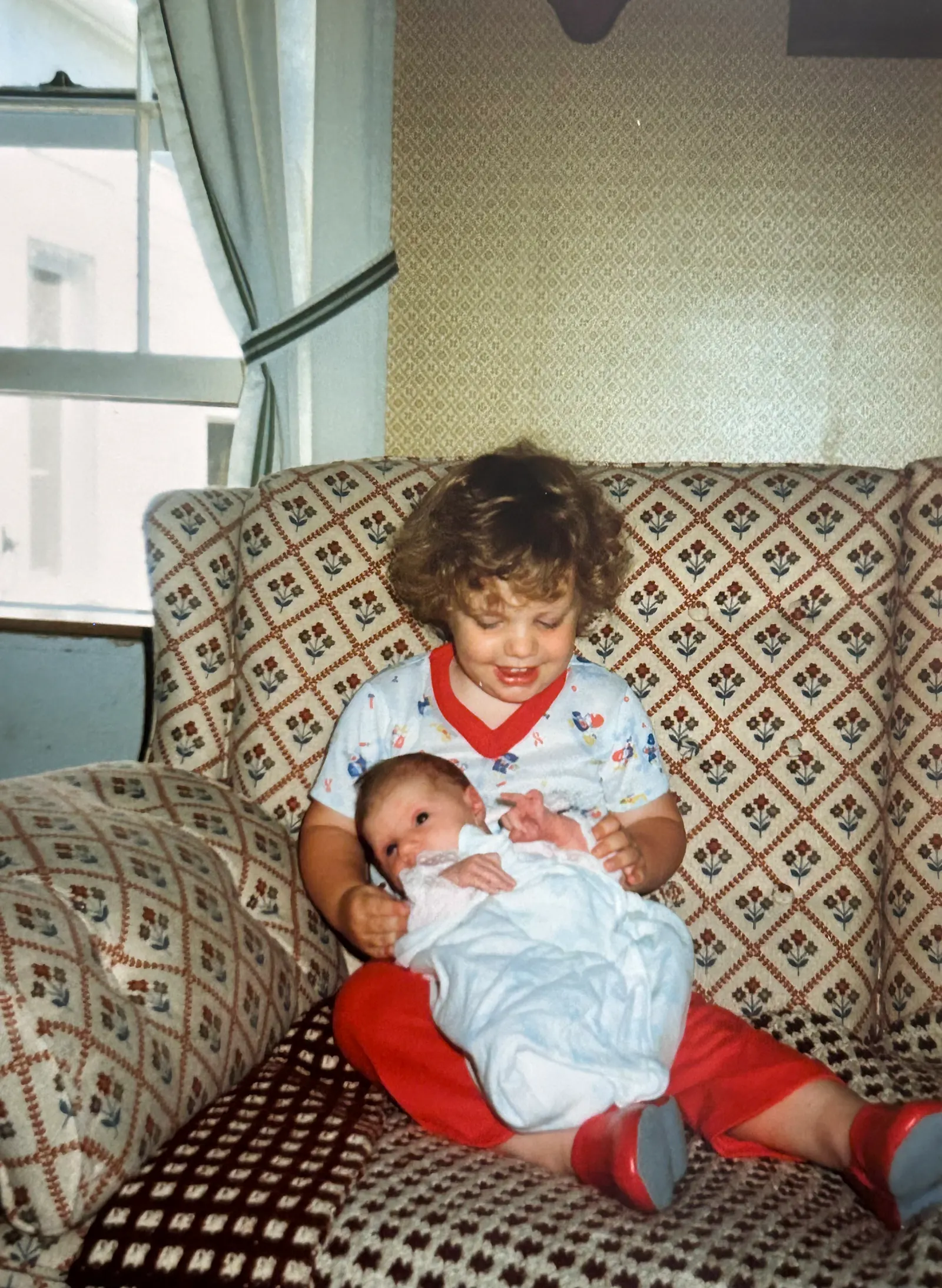 A month old Hannah sitting in my 2-year-old lap. She appears to be trying to sit up while wrapped in a blue blanket. I’m wearing a white outfit with red edges and gaudy red pants. We are sitting on a brown afghan atop a brown couch with dark brown, red, and blue flower patterns. The greenish/brown wallpaper has similar diamond patterns upon it. Behind the couch is a window with white and green curtains. Some style!