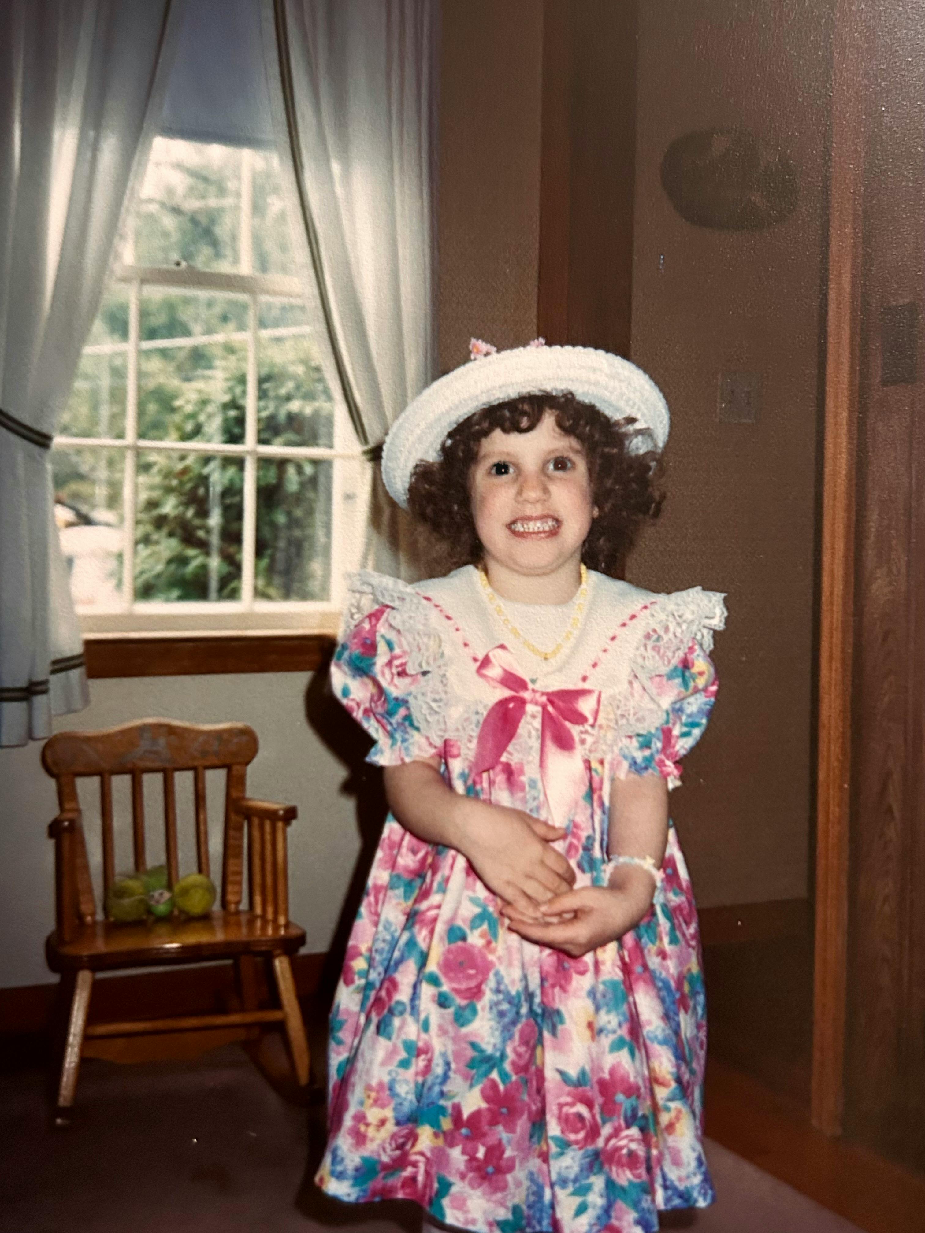 Hannah dressed for Easter in a pink rose dress with blue and white patterns around it. She wears a white bonnet atop her curly hair. A little rocking chair sits behind her beneath a window.