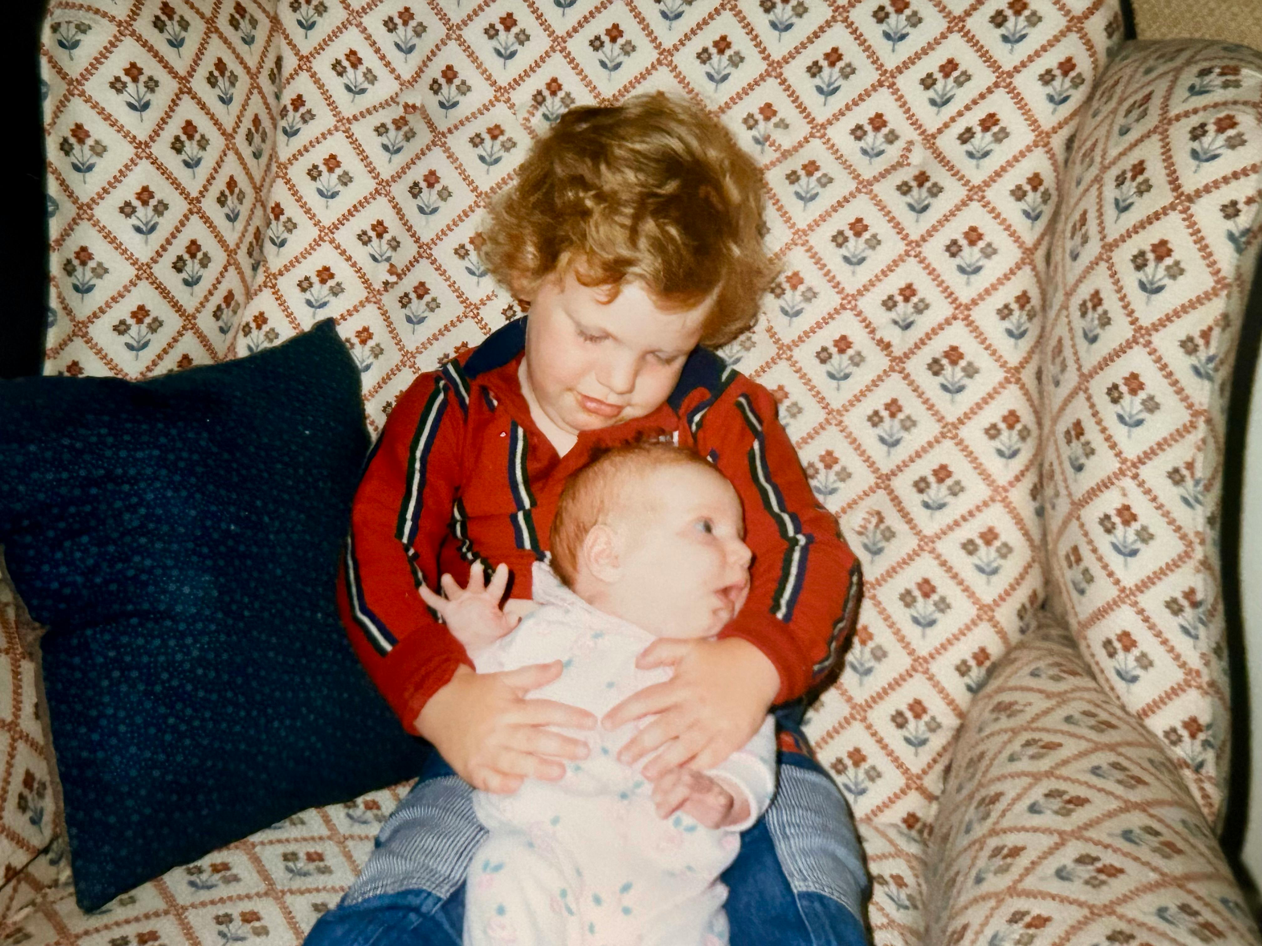 A month old Hannah sitting in my 2-year-old lap. She is looking to the right with a look of surprise on her face as I look down at her. She is wearing a white onesie with little blue flowers on it. I’m wearing a red shirt with black and white vertical stripes. A blue pillow is behind us. We are  sitting on brown couch with dark brown, red, and blue flower patterns.