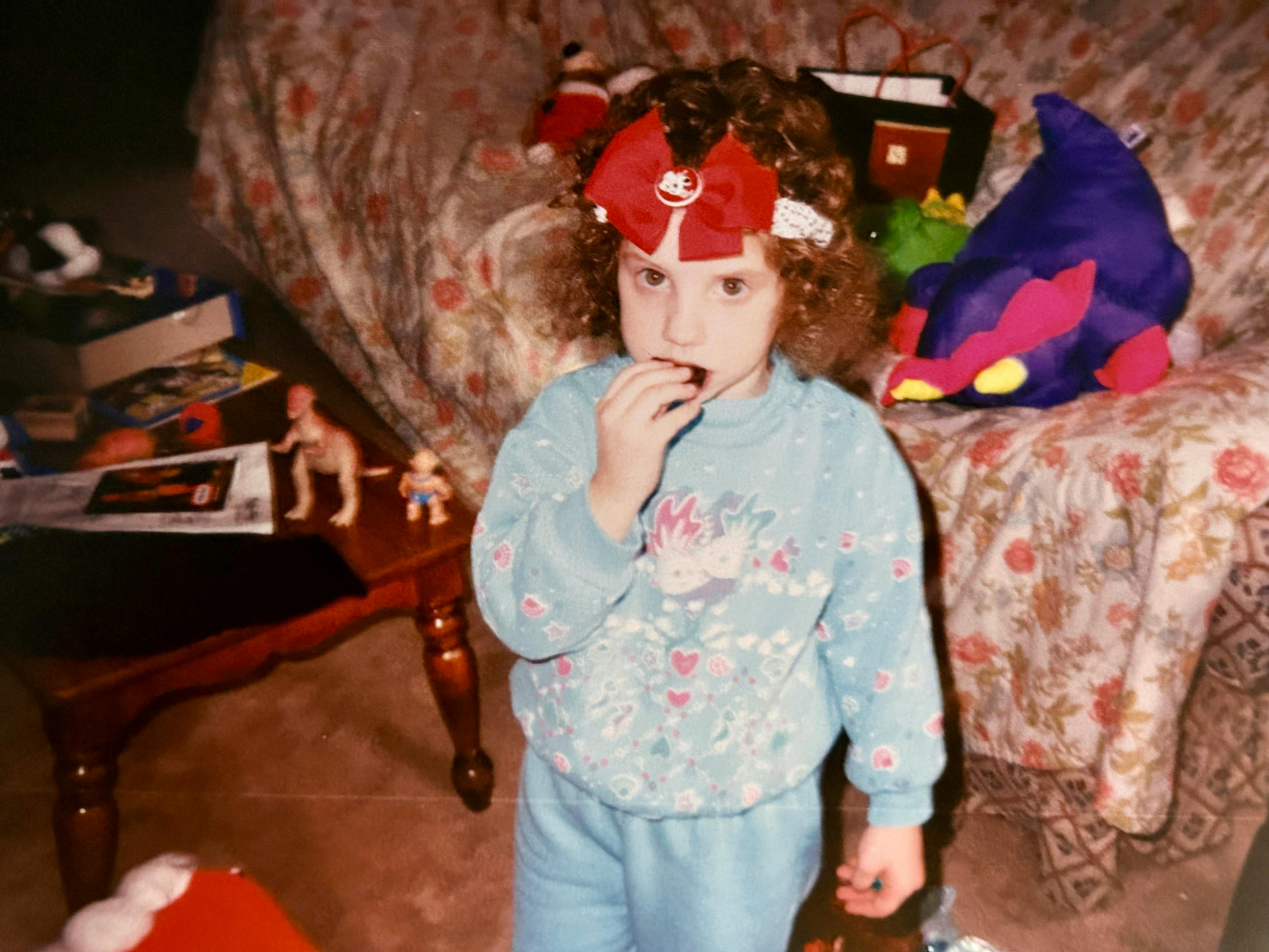 Hannah as a toddler standing before a couch at Christmas with a large red ribbon on her head while she eats a piece of candy. She wears a blue outfit. Various dinosaur and dragon toys fill the room and sit atop the couch covered in a floral patterned throw.