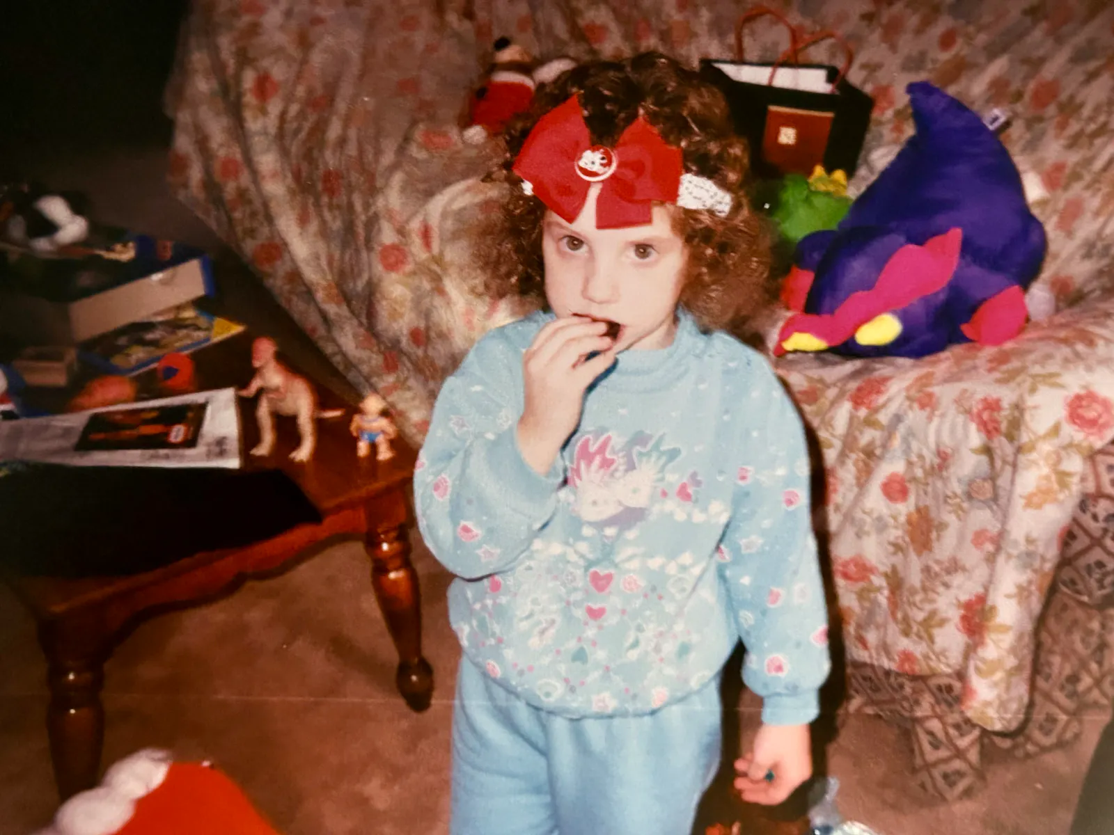 Hannah as a toddler standing before a couch at Christmas with a large red ribbon on her head while she eats a piece of candy. She wears a blue outfit. Various dinosaur and dragon toys fill the room and sit atop the couch covered in a floral patterned throw.