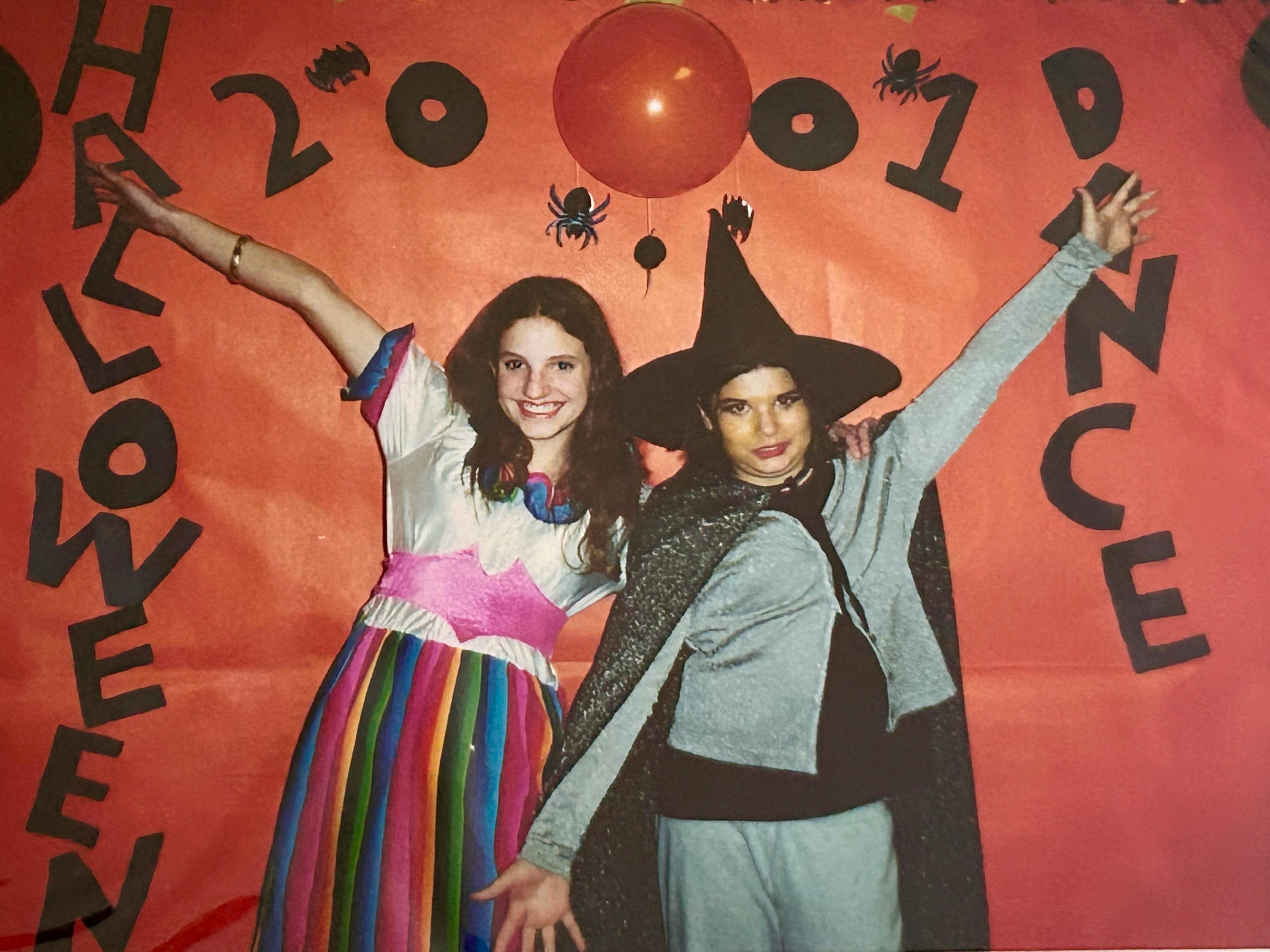 Hannah on the left and her friend Kate on the right before a large orange sign that says “Halloween 2001 Dance” with little drawings of bats and spiders around the text and an orange balloon for one of the 0s in “2001.” Hannah is dressed in a rainbow skirt with a gold bracelet on her right arm, her friend Kate is dressed as a stereotypical witch with a grey costume, black cape, and black pointed hat.