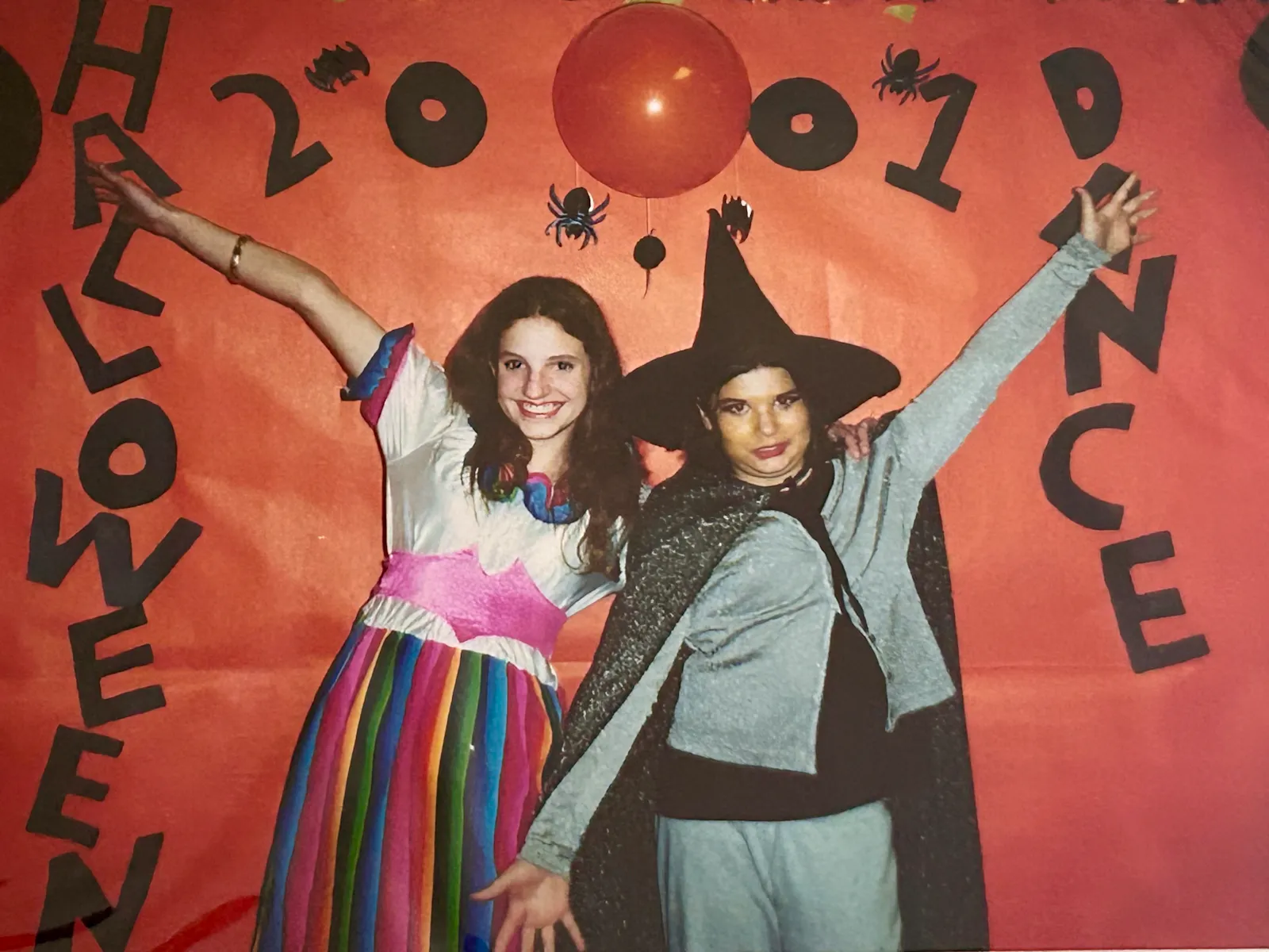 Hannah on the left and her friend Kate on the right before a large orange sign that says “Halloween 2001 Dance” with little drawings of bats and spiders around the text and an orange balloon for one of the 0s in “2001.” Hannah is dressed in a rainbow skirt with a gold bracelet on her right arm, her friend Kate is dressed as a stereotypical witch with a grey costume, black cape, and black pointed hat.