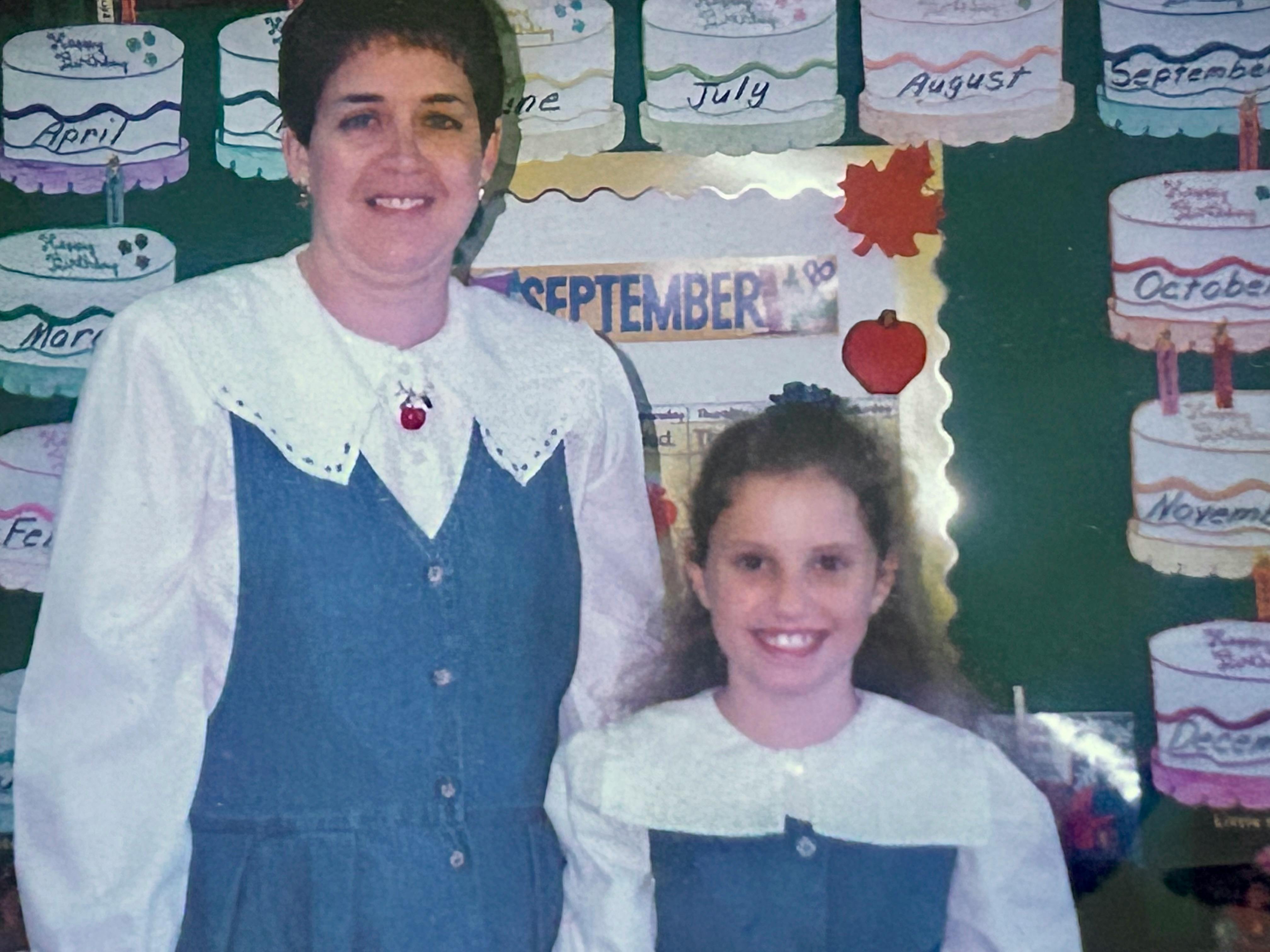 Hannah in the First Grade with her teacher, both wearing the same outfit: a blue denim blouse with white edges and sleeves. They stand in front of a board covered in hand drawn cakes with the months of the year written on them and “Happy Birthday” on to tops where the students would have their names displayed in their birthday months. The calendar behind them shows that it was taken in September.