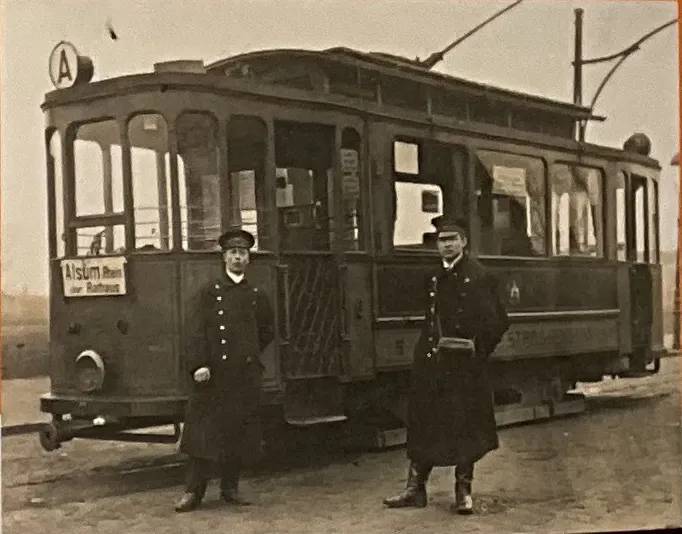 Schwarzweiß-Foto. Das Bild wir ausgefüllt von dem Triebwagen 23, einer alten Straßenbahn. Davor posieren zwei Männer in Schaffner-Uniform