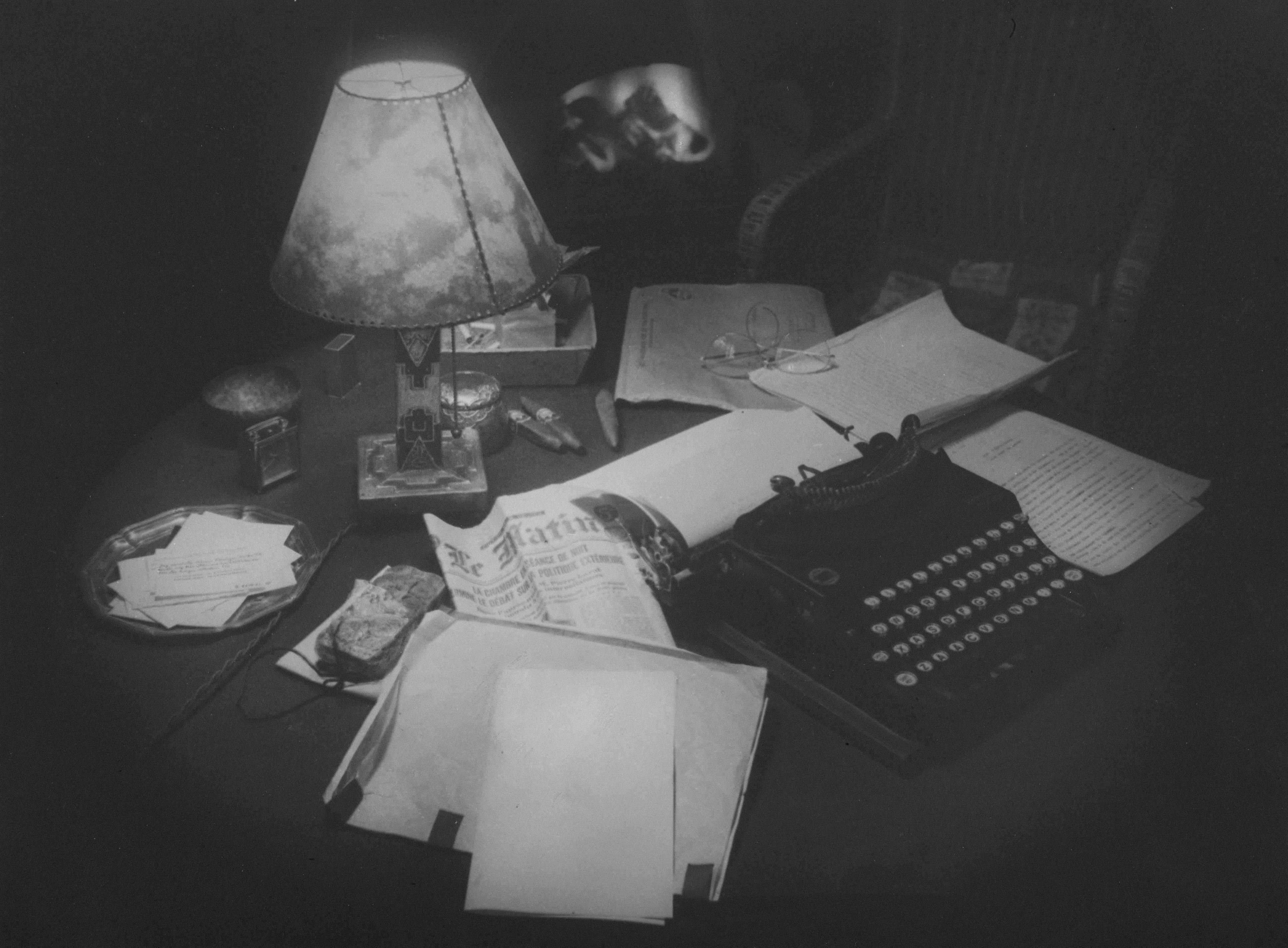 Schwarz-weiß Bild von einem Schreibtisch mit alter Lampe, alter Schreibmaschine und jede Menge Papierkram.