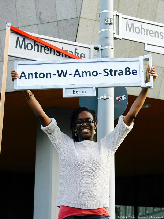 Sharon hält ein neues Straßenschild mit der Aufschrift „Anton-W-Amo-Straße“ hoch. Hinter ihr ist der alte Name zu sehen, der mit einem roten Strich durchgestrichen ist.