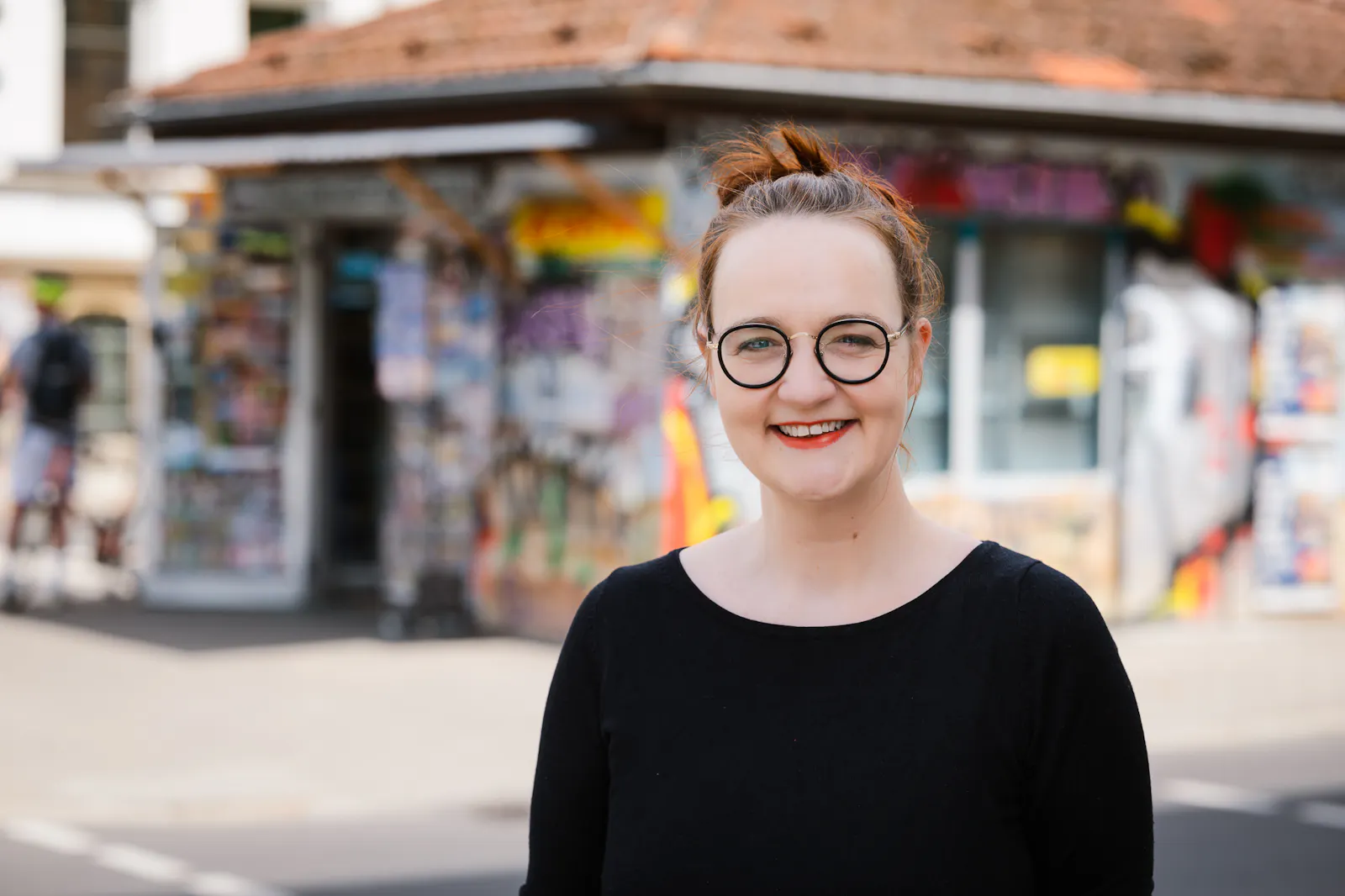 Insa van den Berg, Frau mit roten Haaren und Brille, vor Zeitungskiosk.