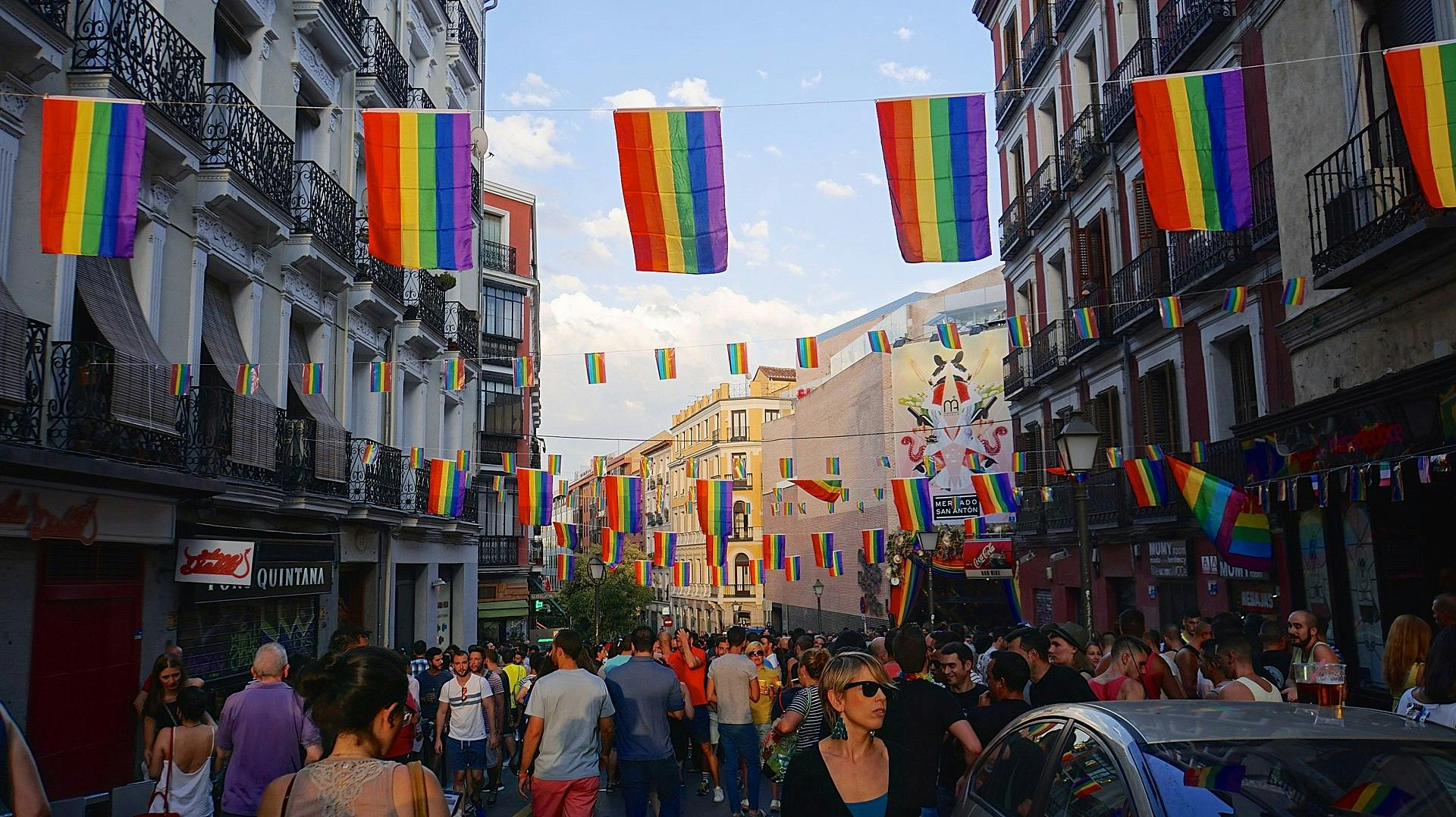 Eine belebte Straße in Madrid, zwischen den Häusern sind Girlanden mit Regenbohnenfahnen gespannt.