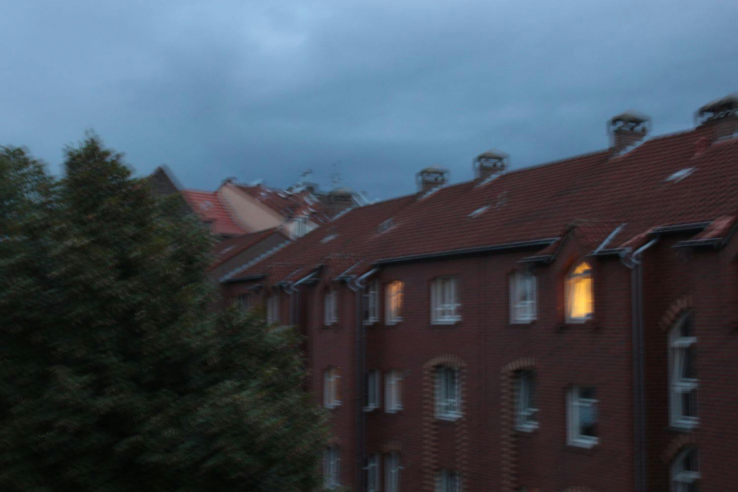Verschwommene Aussicht auf ein rotes Wohnhaus, in einigen Fenstern brennt Licht. Der Himmel ist bedrohlich grau-blau.