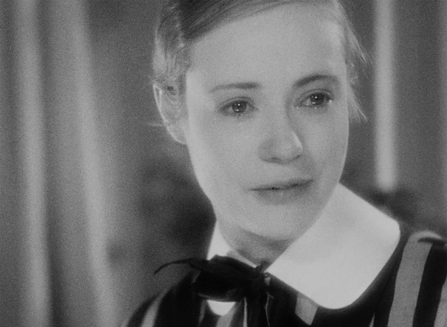 Nahaufnahme in Schwarz-Weiß der Manuela von Meinhardis (gespielt von Hertha Thiele) aus dem Film Mädchen in Uniform (1931). Die Figur hat Tränen in den Augen und blickt niedergeschlagen nach unten. Sie trägt eine dunkle Uniform mit breiten, hellen Schulterstreifen, einem großen, weißen Peter-Pan-Kragen und einer dunklen Schleife am Hals. Das Haar ist streng zurückgekämmt, und die Beleuchtung ist weich, was die emotionale Intensität unterstreicht.