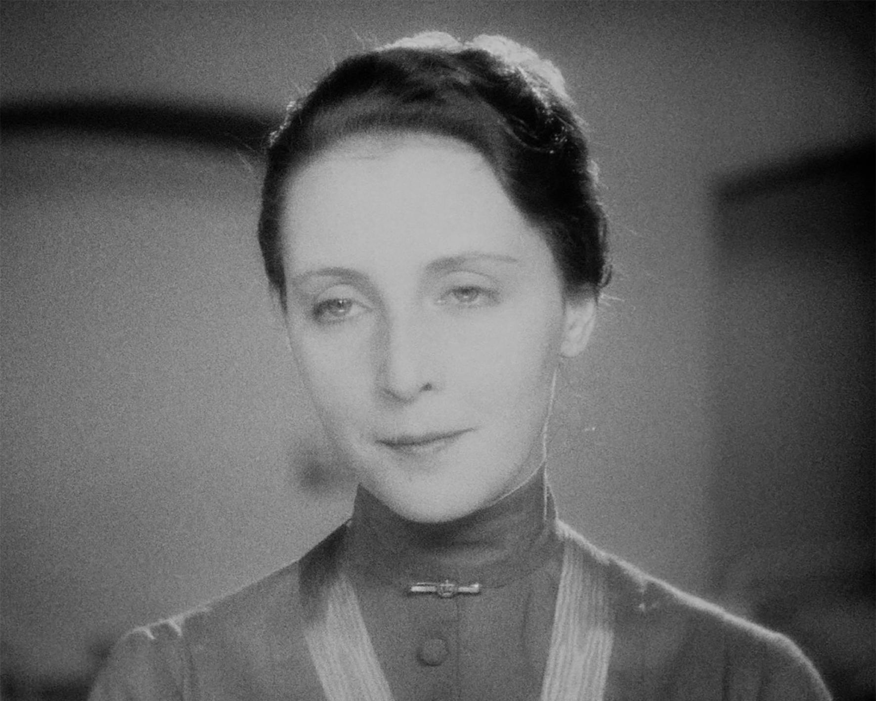 Nahaufnahme in Schwarz-Weiß der Fräulein von Bernburg (gespielt von Dorothea Wieck) aus dem Film Mädchen in Uniform (1931). Die Darstellerin blickt mit einem sanften, leicht melancholischen Ausdruck nach vorne. Sie trägt ein hochgeschlossenes, dunkles Kleid oder eine Bluse mit einem kleinen, verzierten Broschen-Verschluss am Kragen. Ihr dunkles Haar ist elegant zurückgesteckt. Die Beleuchtung ist weich, konzentriert sich auf ihr Gesicht und erzeugt einen nachdenklichen, würdevollen Eindruck.