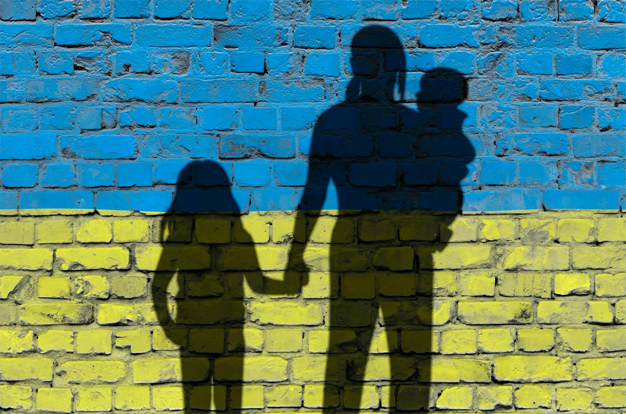 Das Bild zeigt die Schatten einer Familie mit zwei Kindern auf einer Ziegelmauer, die in den Farben der ukrainischen Flagge bemalt ist: oben blau, unten gelb.