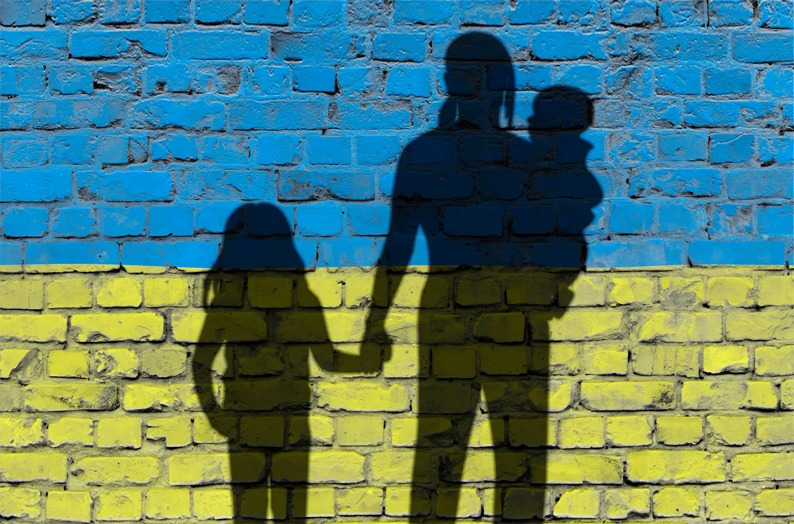 Das Bild zeigt die Schatten einer Familie mit zwei Kindern auf einer Ziegelmauer, die in den Farben der ukrainischen Flagge bemalt ist: oben blau, unten gelb.