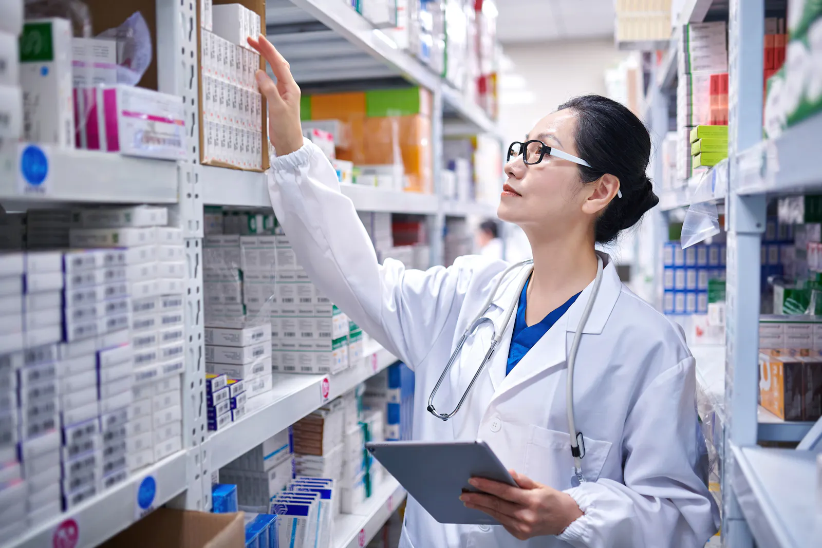 Das Bild zeigt eine Apothekerin im weißen Kittel. Sie greift nach einer Medikamenten-Packung in einem Apotheken-Regal. In der anderen Hand hält sie ein Tablet.