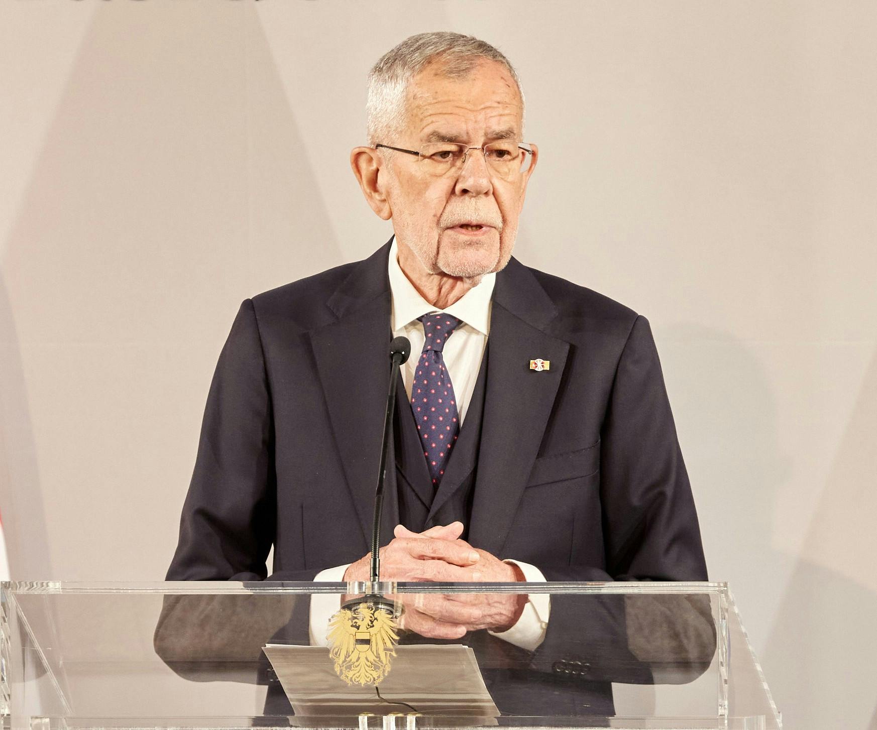 Das Bild zeigt Bundespräsident Alexander Van der Bellen. Er steht an einem Rednerpult aus Glas mit dem österreichischen Bundesadler. Er trägt einen dunklen Anzug mit blauer Krawatte.