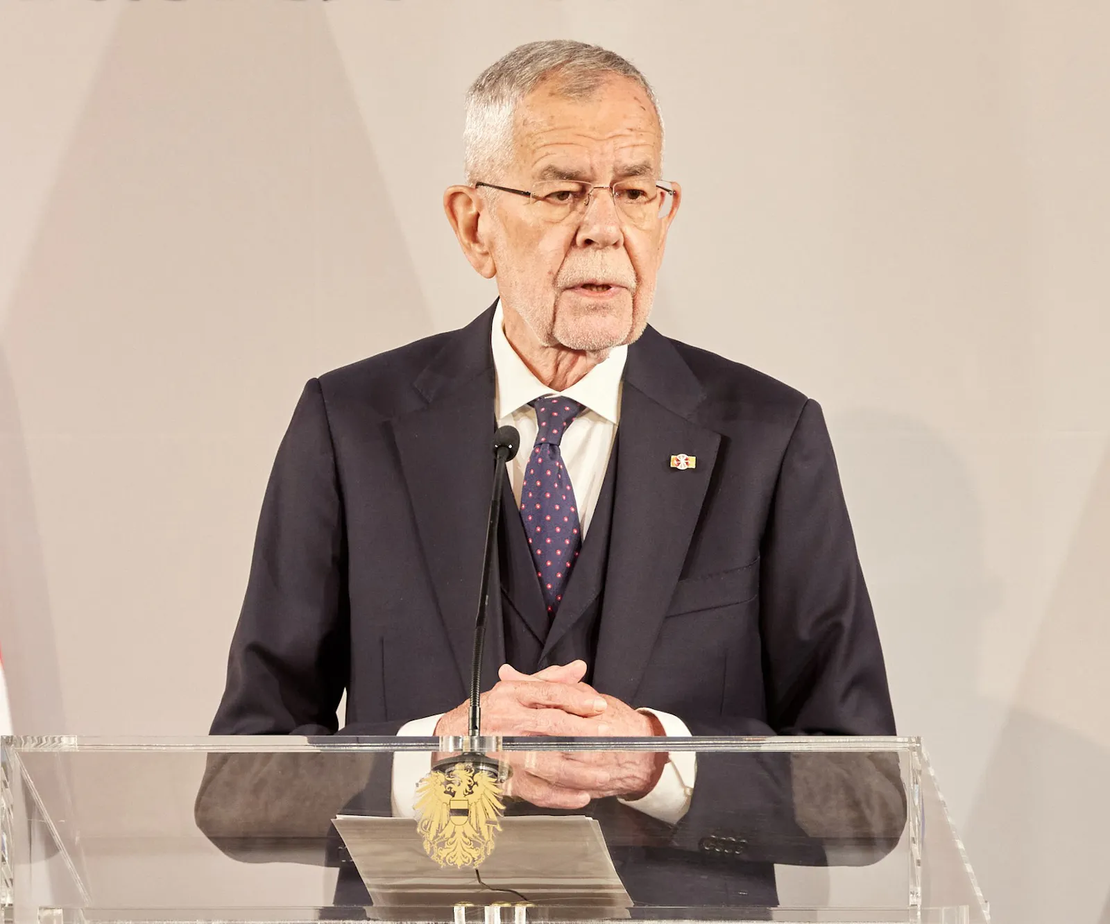 Das Bild zeigt Bundespräsident Alexander Van der Bellen. Er steht an einem Rednerpult aus Glas mit dem österreichischen Bundesadler. Er trägt einen dunklen Anzug mit blauer Krawatte.