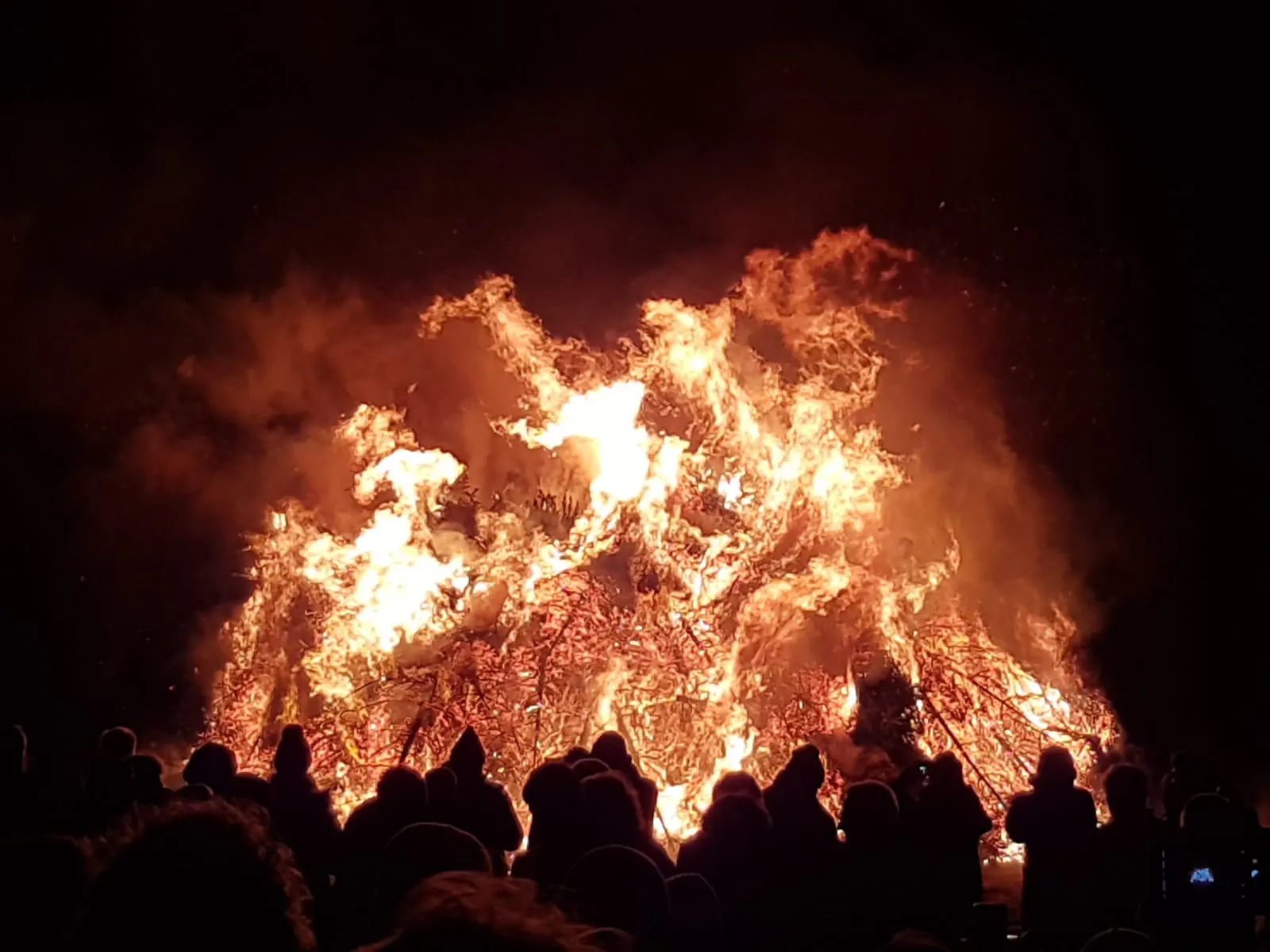 Biikefeuer. Im Vordergrund Menschen, die sich als schwarze Silhouetten vor den leuchtenden Flammen abheben.