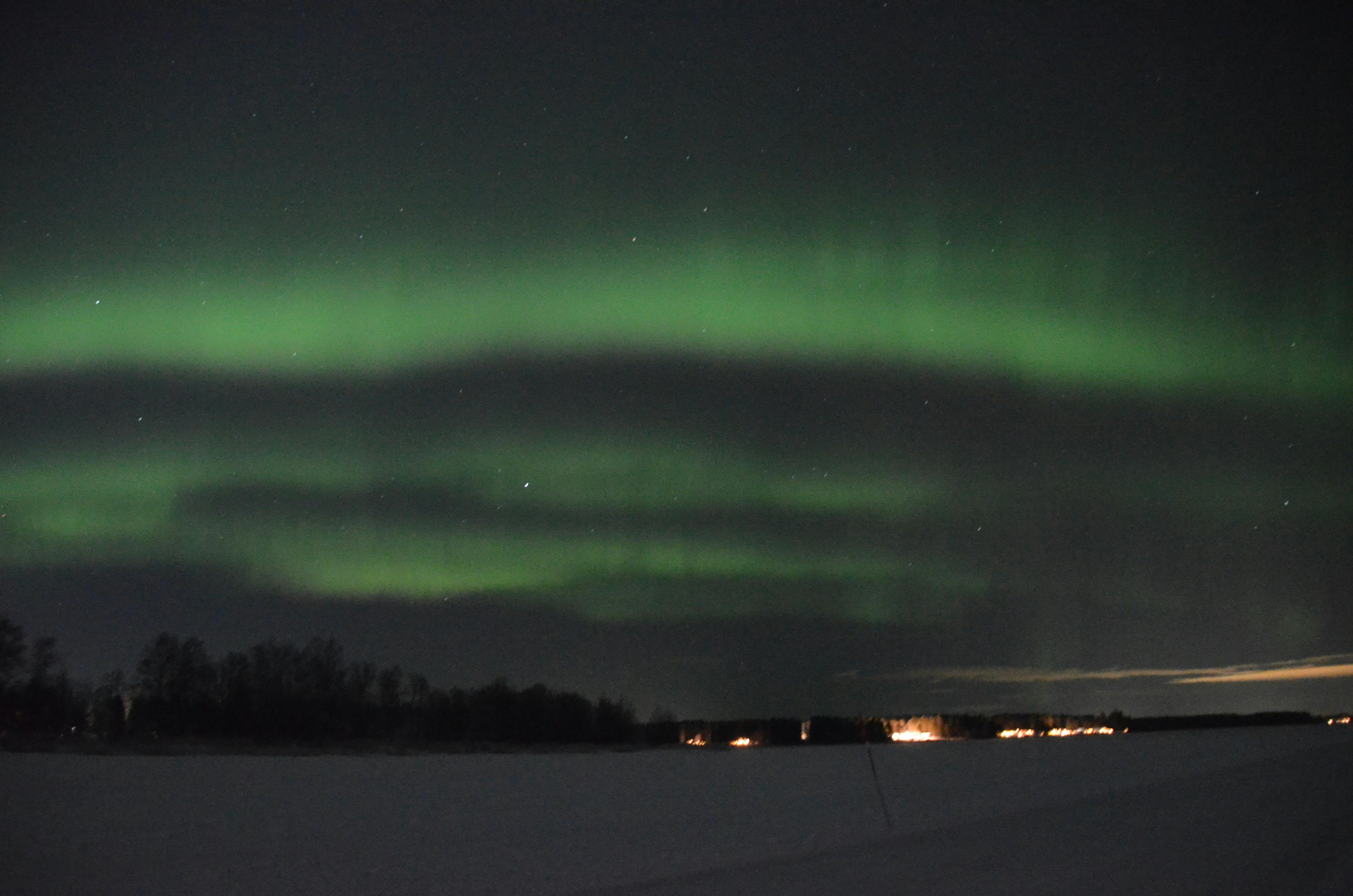 Nacht, grüne Streifen am Himmel, unten schneebedeckt, im Hintergrund Lichter