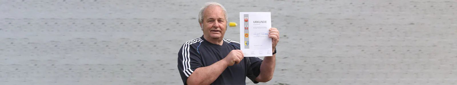 Ein älterer Mann steht vor einem grauen Hintergrund. Er trägt Sportbekleidung und hält eine Teilnehmerurkunde hoch.